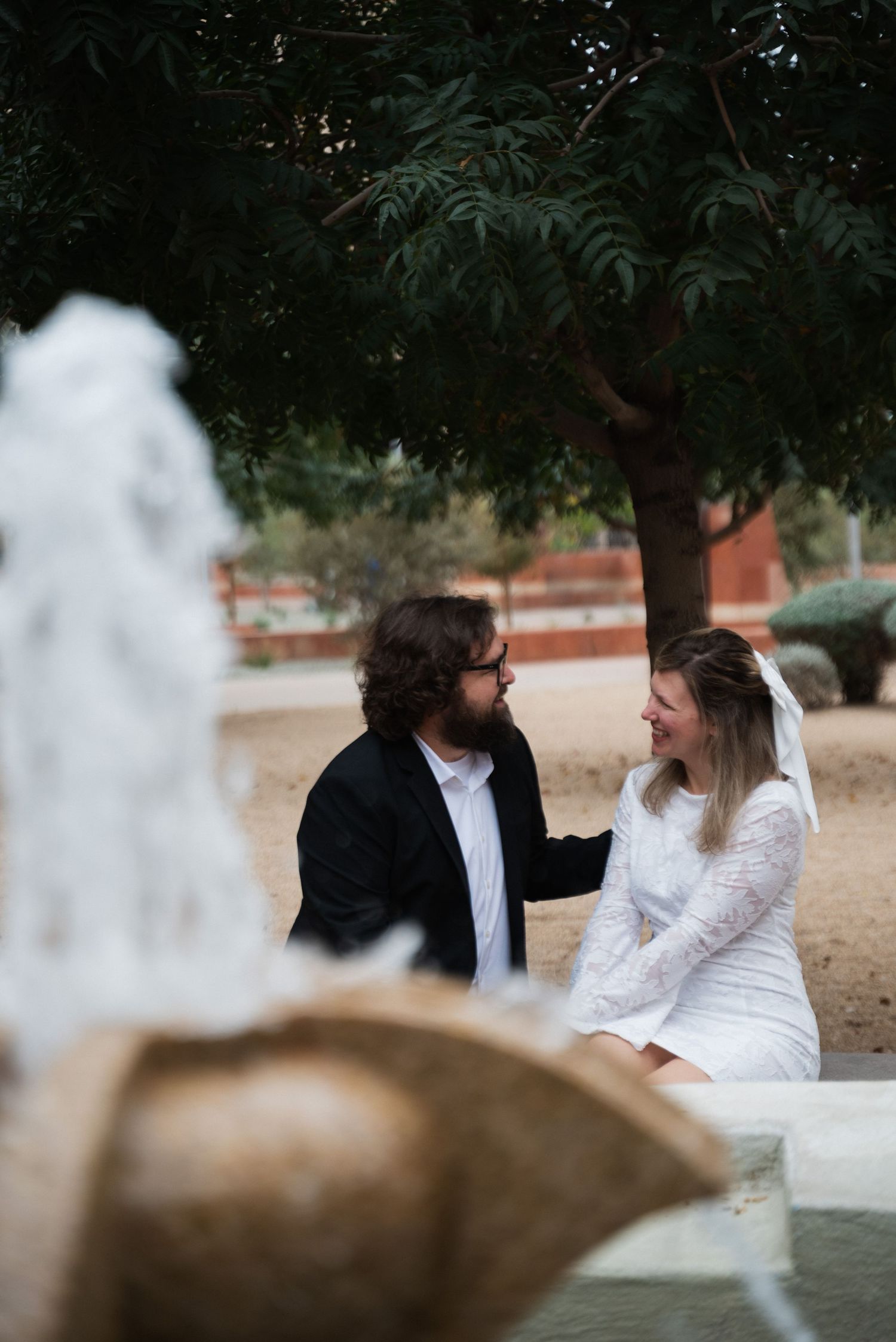 A couple in wedding attire sits together on a stone bench in a garden setting during their photoshoot.