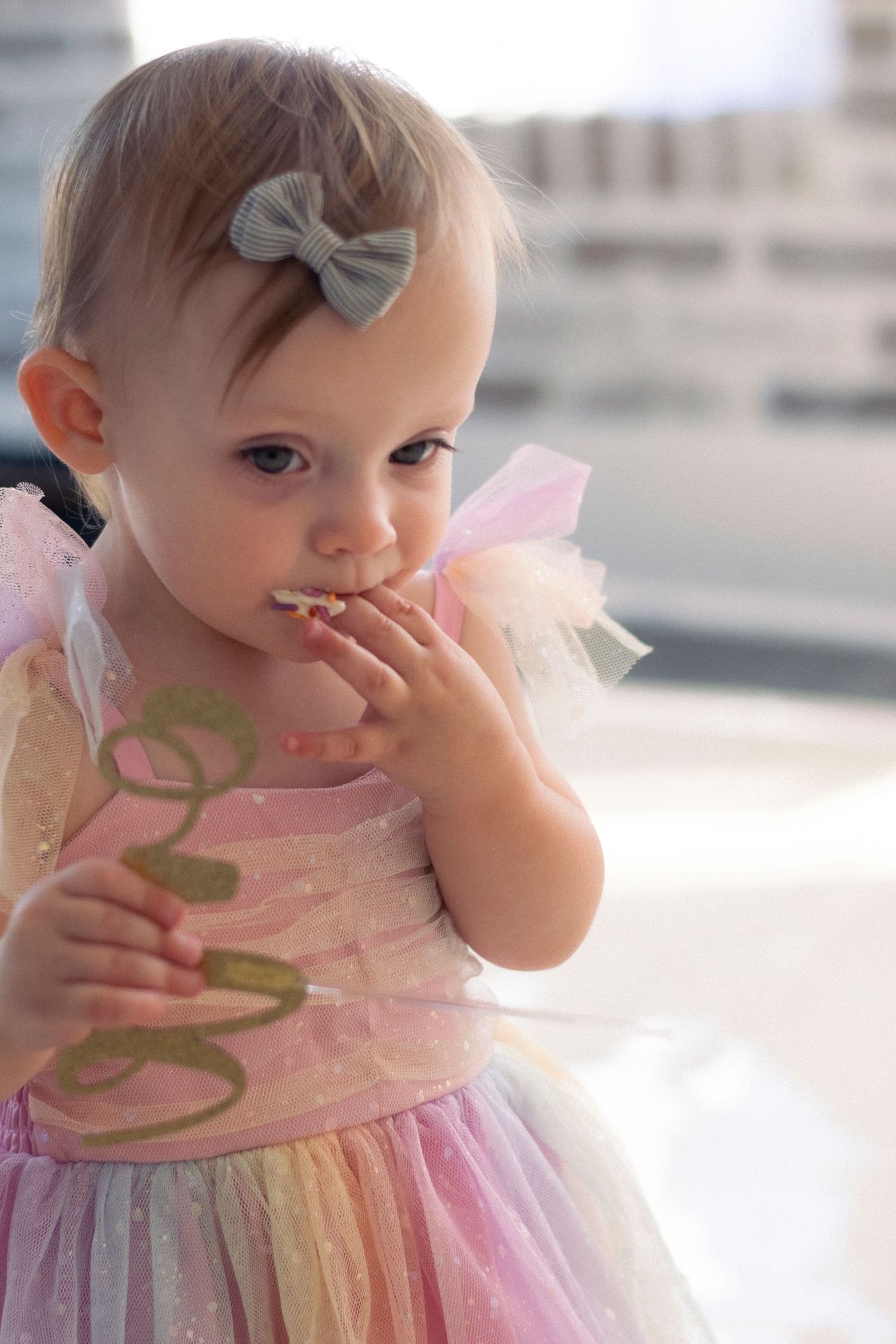 Baby in a pink tulle dress with a gray bow wearing pastel colors stands contemplatively with hand near mouth.