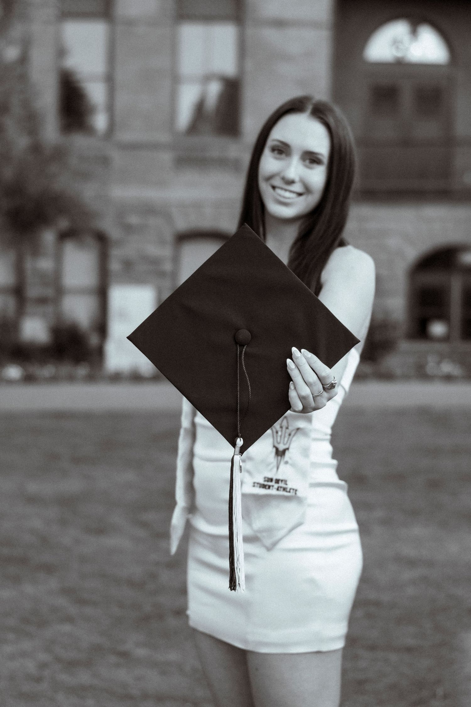A graduate in a white dress holds their graduation cap against an architectural background in black and white.