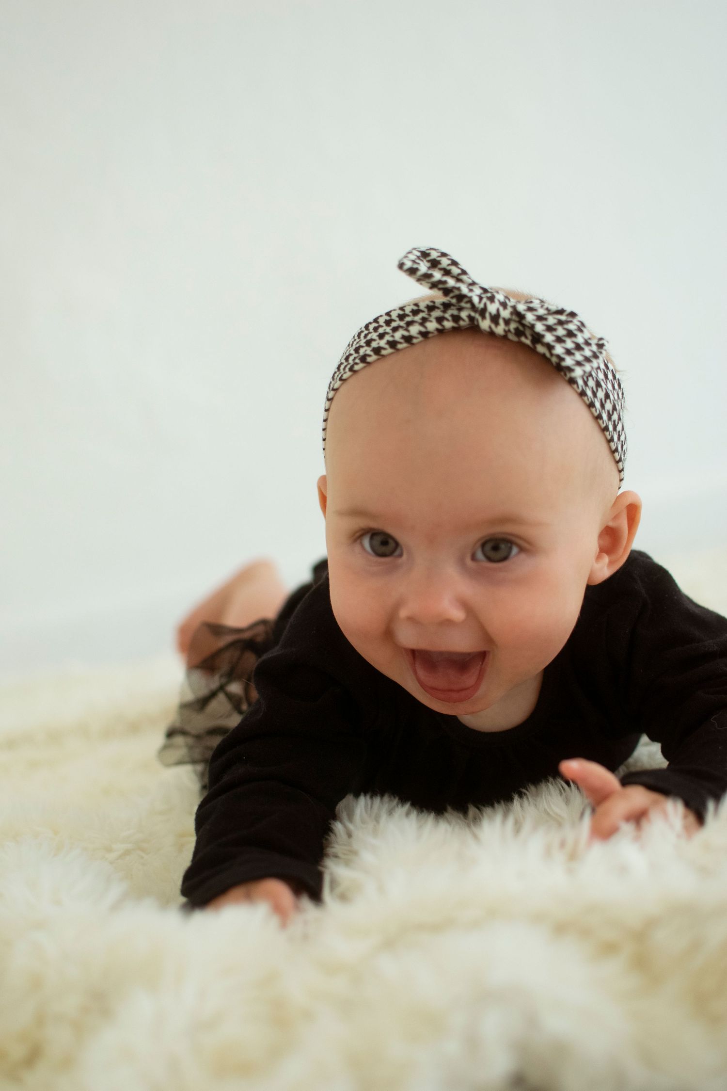 Baby wearing black top and checkered headband lies on fluffy white rug while smiling at camera.