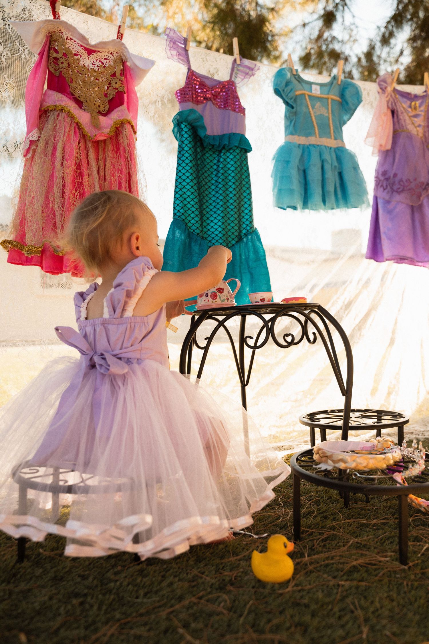 Toddler girl in lavender tutu dress reaches toward princess costumes hanging outdoors at a fairy tale themed birthday party.