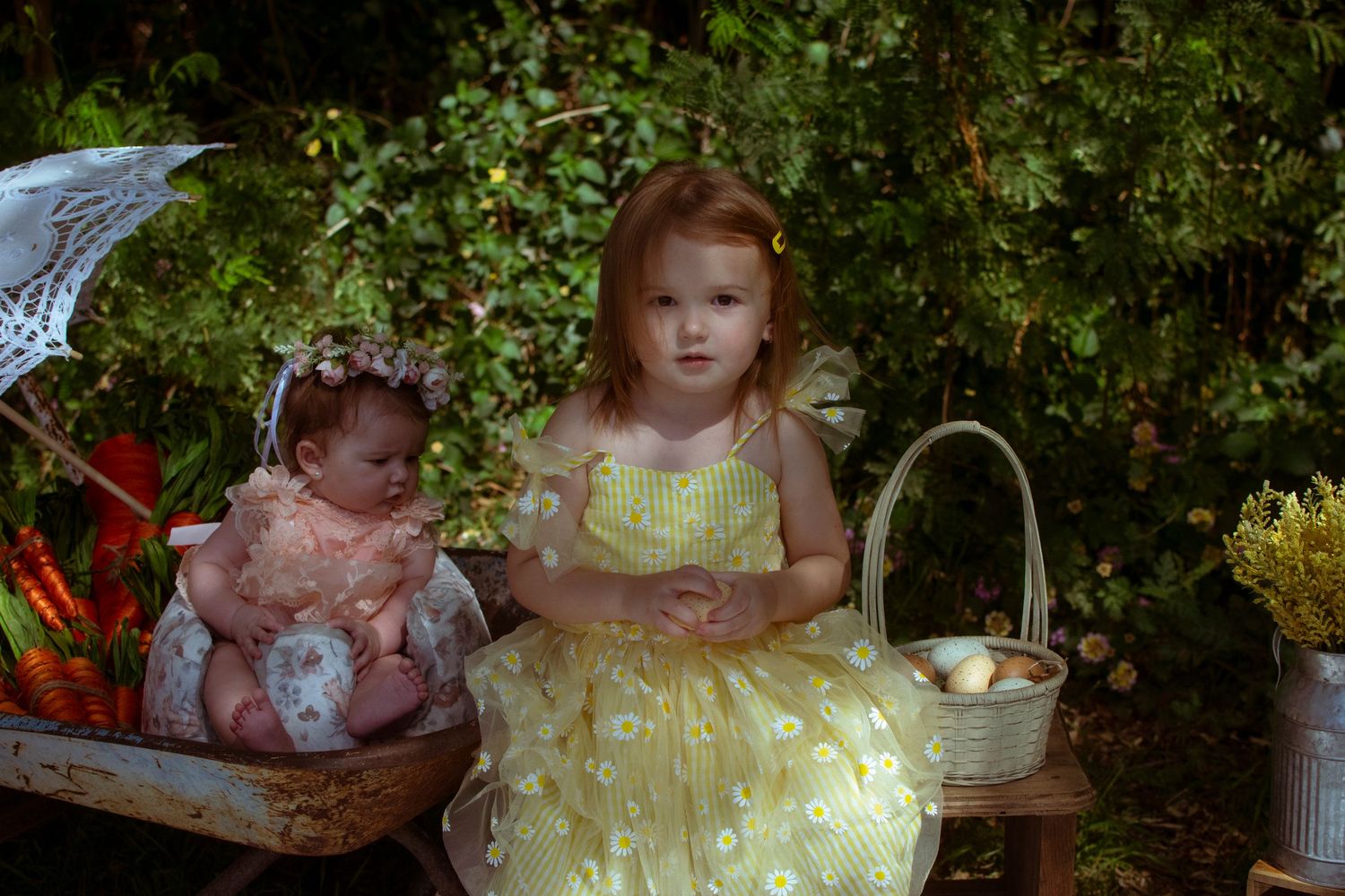 Two young children in fancy dresses sit outdoors with decorative baskets and floral arrangements.