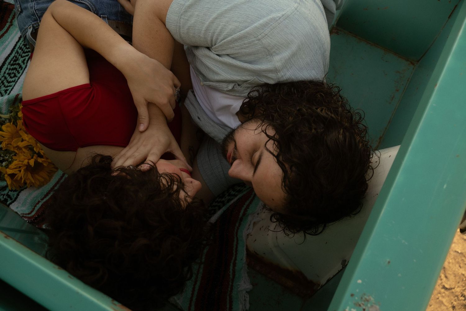 Two young women with curly hair lying close together on a teal surface, sharing a tender moment.