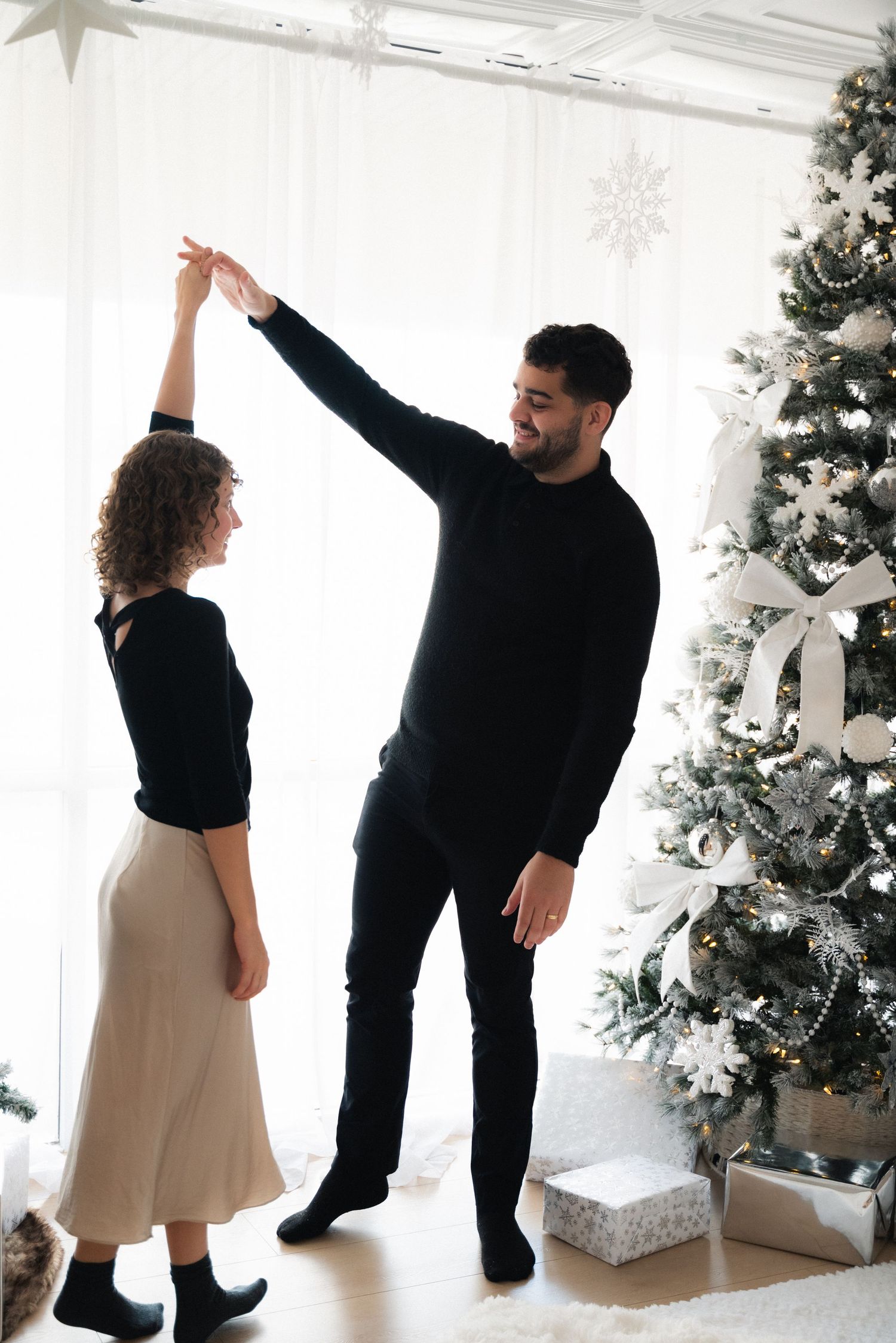 A couple in black and white outfits dances playfully near a decorated Christmas tree in a bright white room.