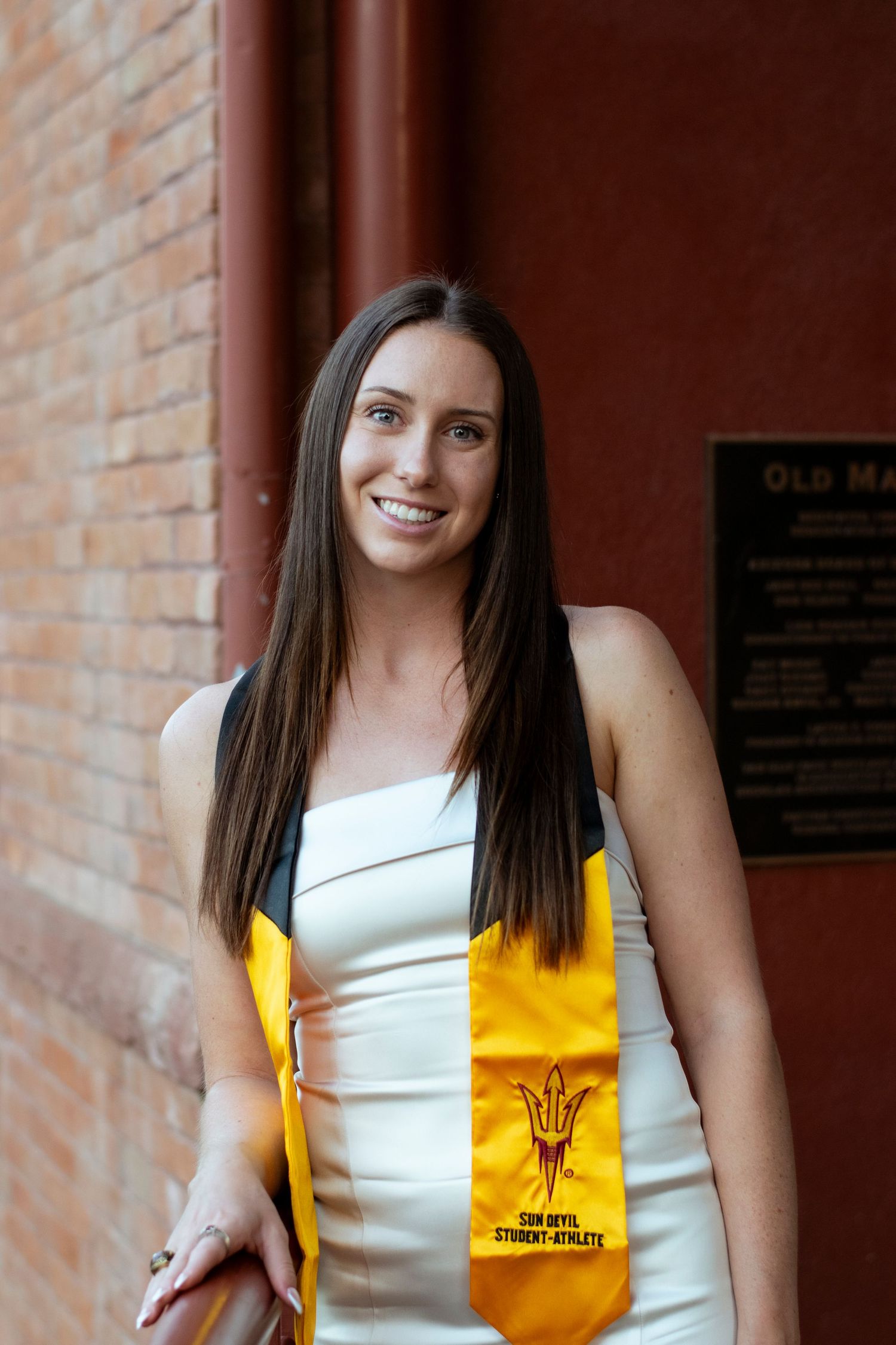 Student wearing a white dress and yellow graduation stole smiles in front of brick wall at university.