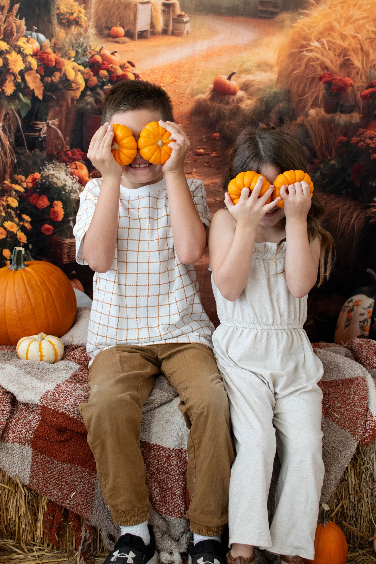 Two children holding small pumpkins over their eyes sit on hay bales in fall setting.