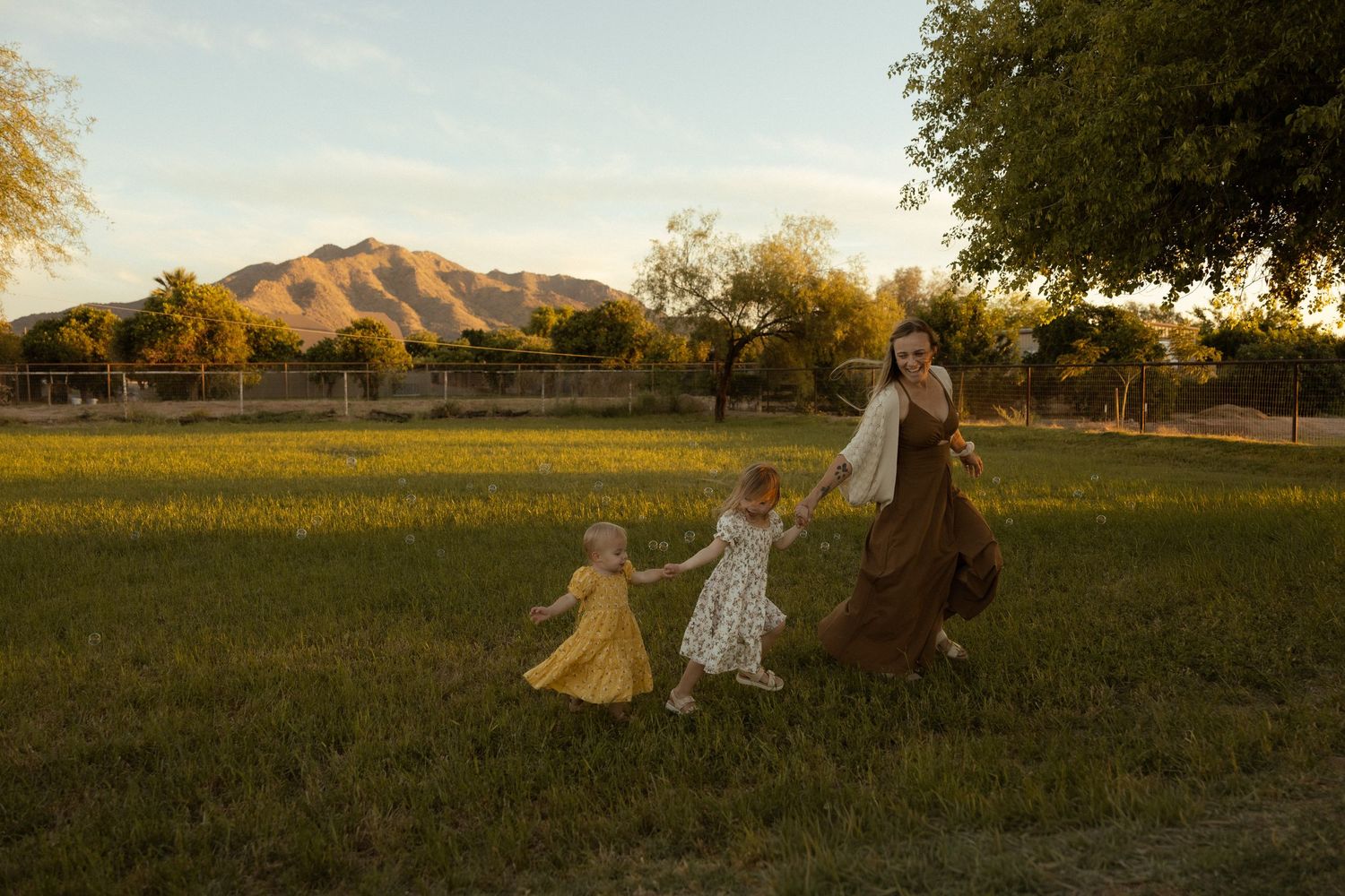 Mother and two young daughters hold hands dancing in a sunlit green field with a mountain backdrop at golden hour.