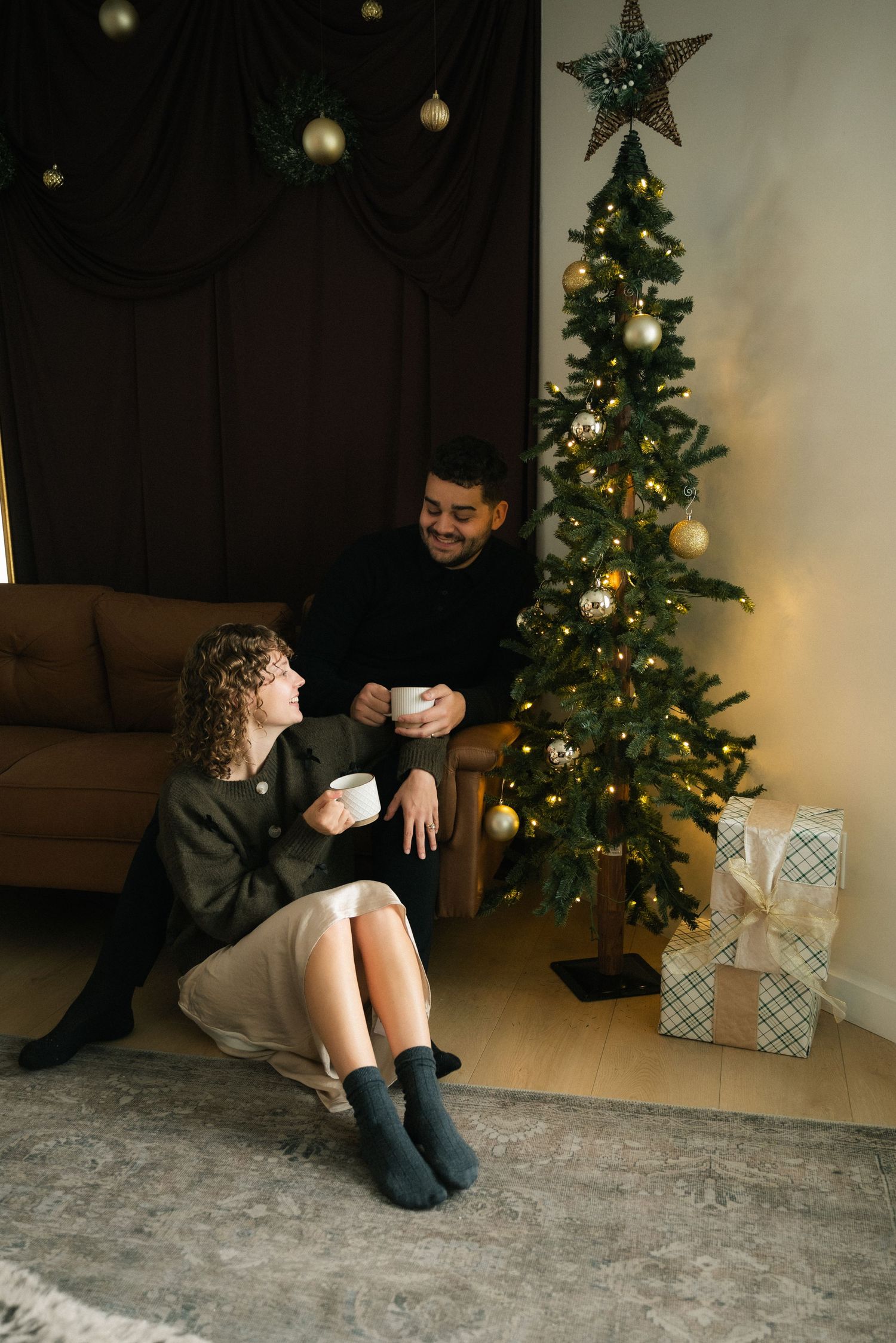 A cozy Christmas scene with a decorated tree, wrapped presents, and two people sitting on a grey carpet.