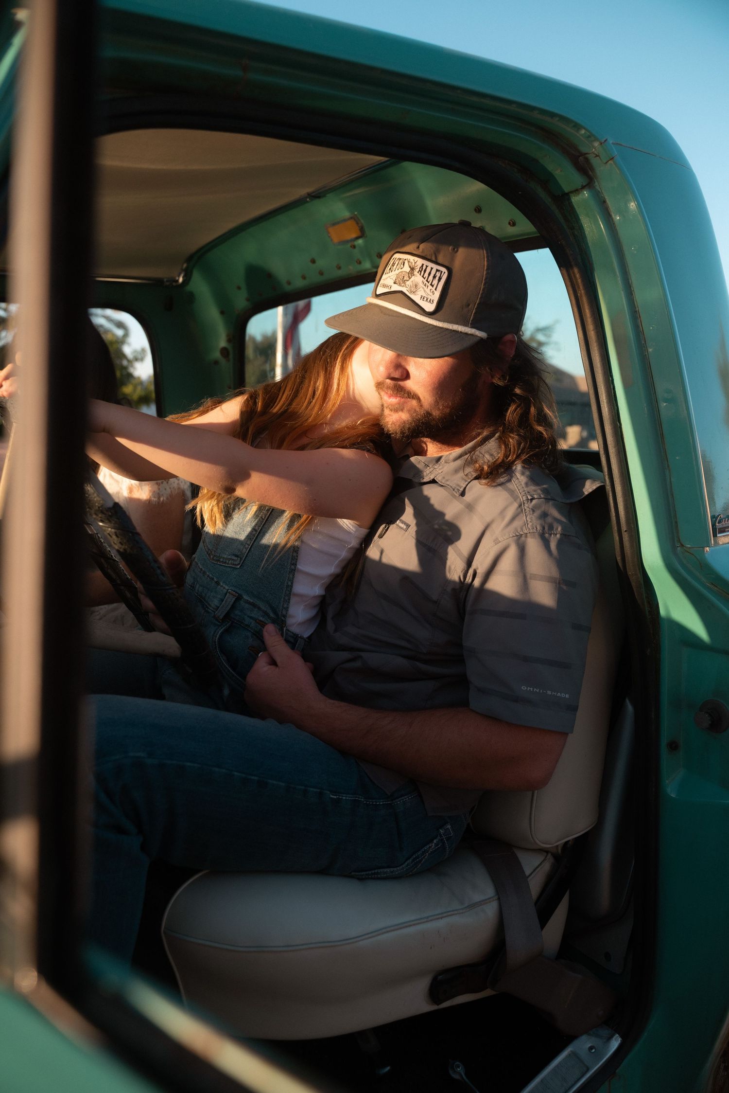 Couple sharing a romantic moment in a vintage teal truck, man in gray shirt and cap, woman in denim overalls.