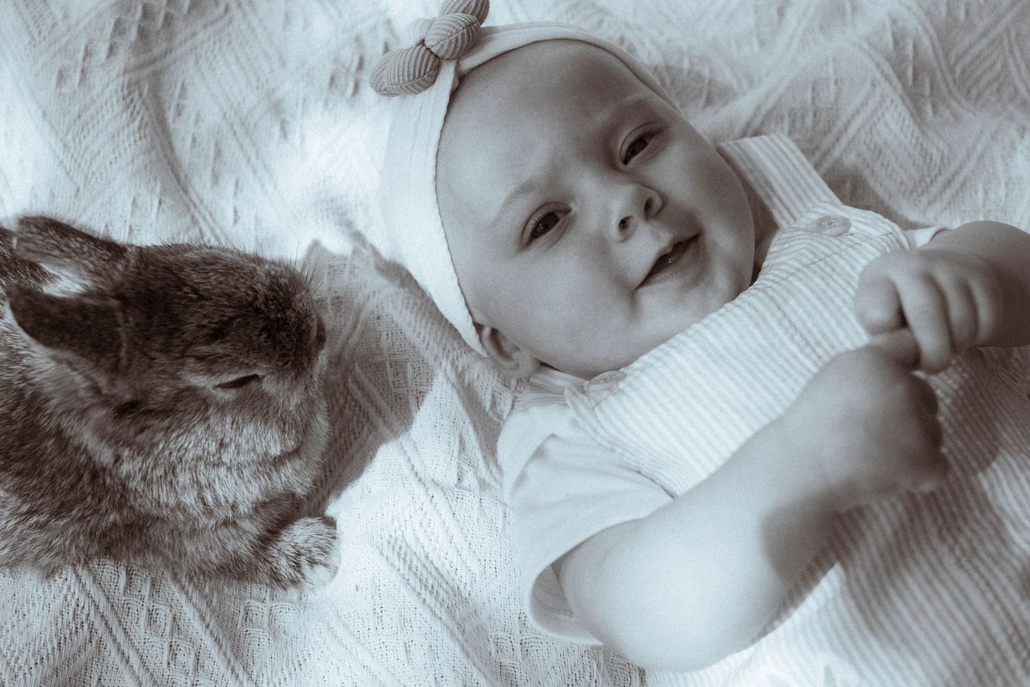 A young infant in white clothing lying next to a bunny rabbit on light colored fabric.