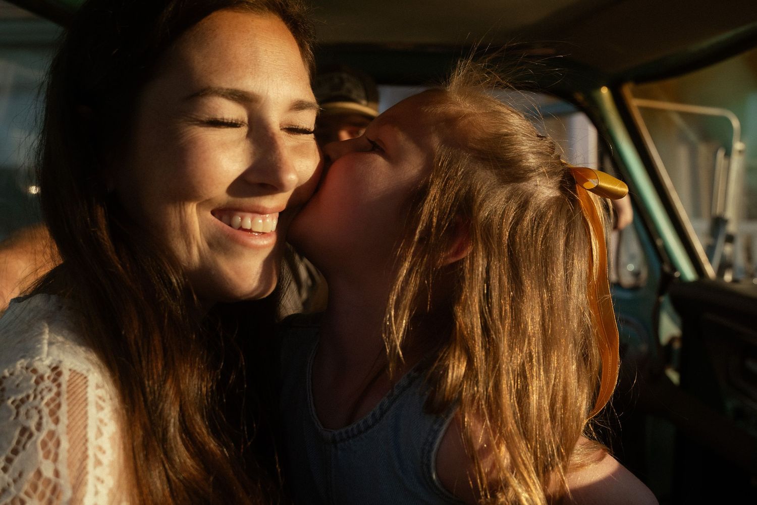 A young girl kisses her smiling mother on the cheek inside a vintage car in warm golden sunlight.