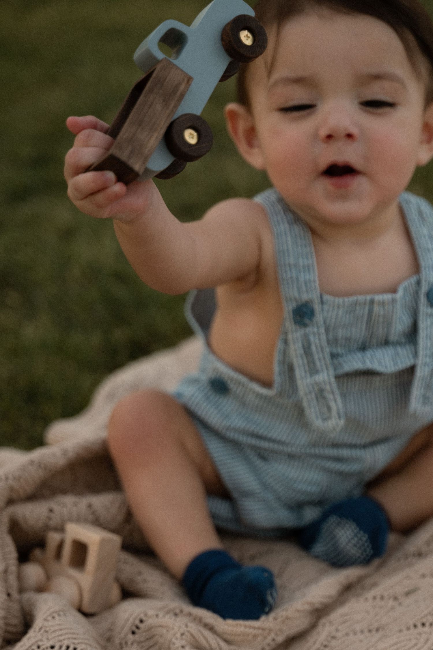 Toddler in blue romper sitting on knit blanket outdoors, holding wooden toy car up while playing.