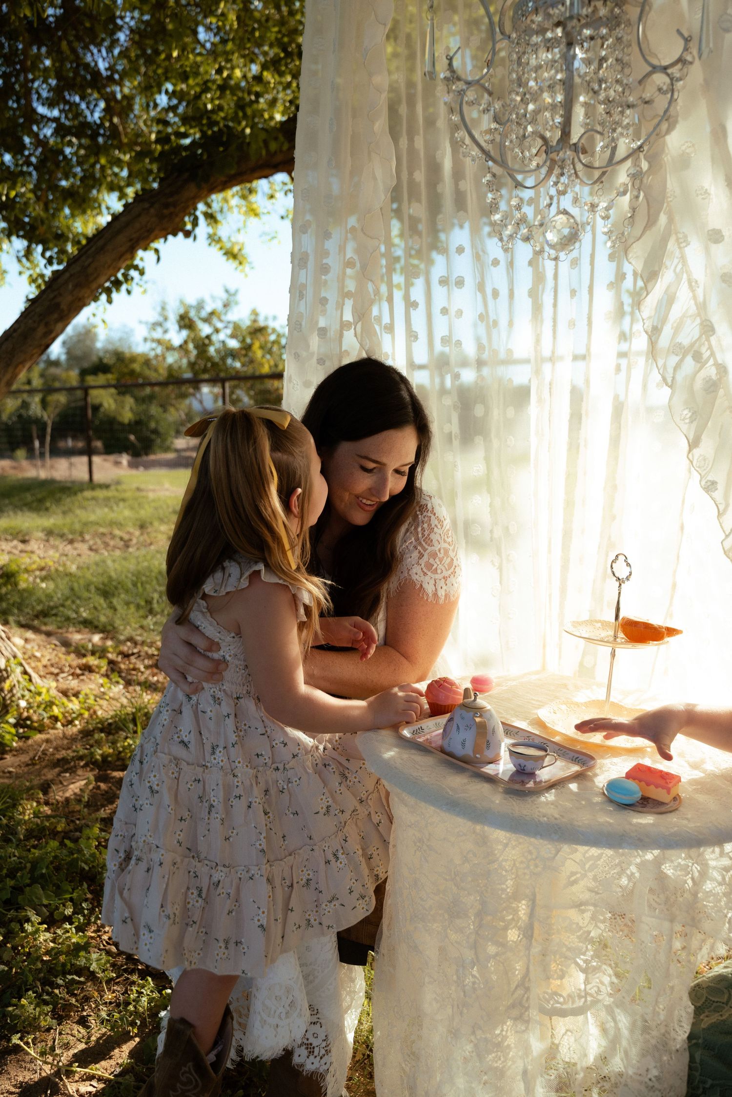 A mother and young daughter share a tender moment at an outdoor lace-decorated tea party table.
