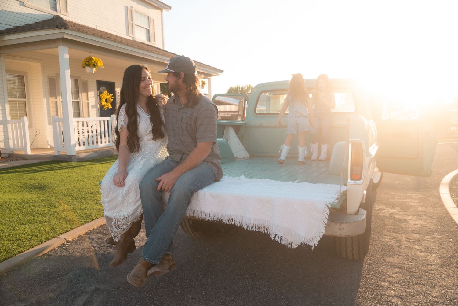 Couple sitting on tailgate of white truck at sunset outside farmhouse home.
