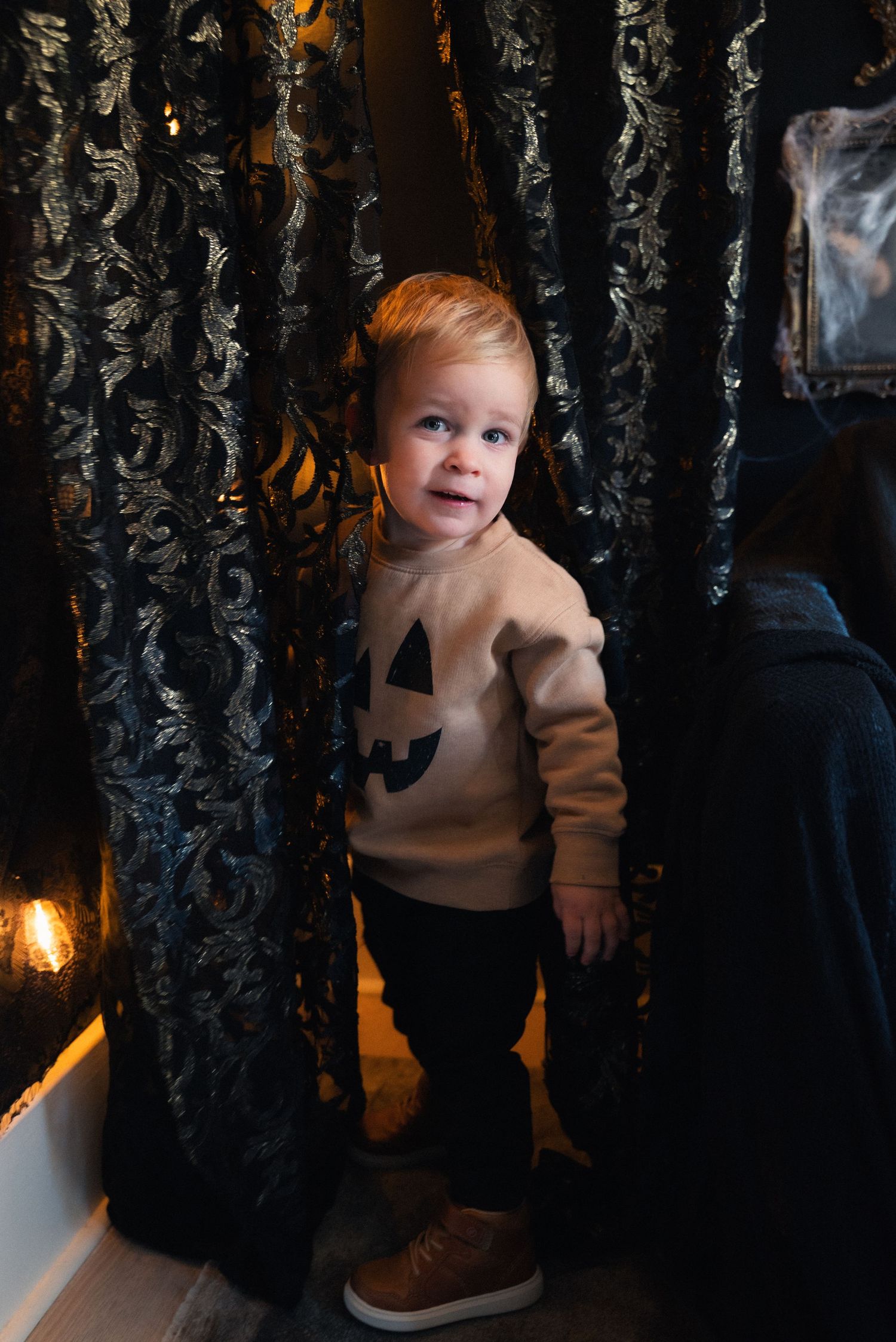 A small child in a beige sweater explores a dark space with ornate black curtains and warm lighting.