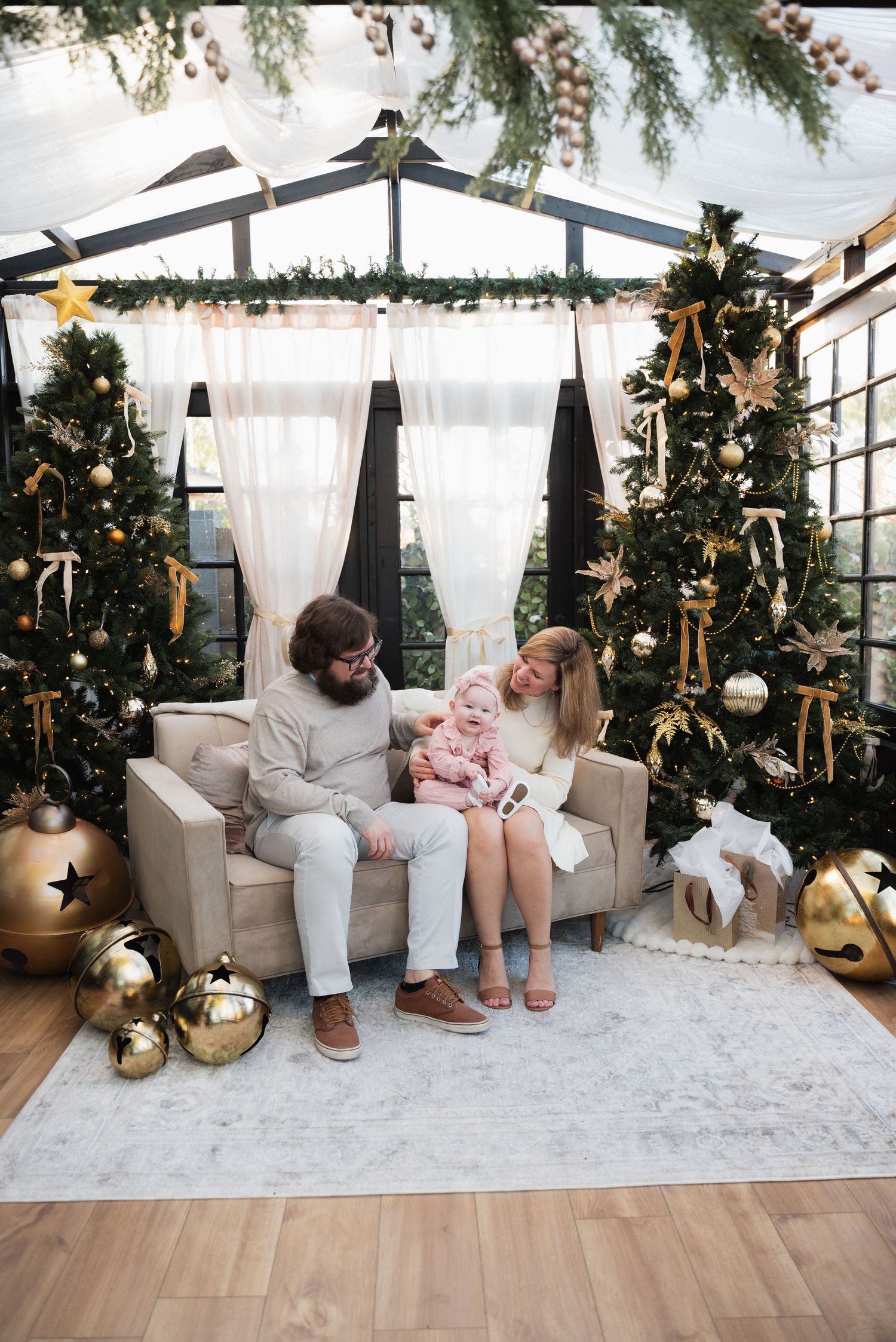 Family with baby sitting on couch surrounded by decorated Christmas trees and gold ornaments in festive indoor setting.