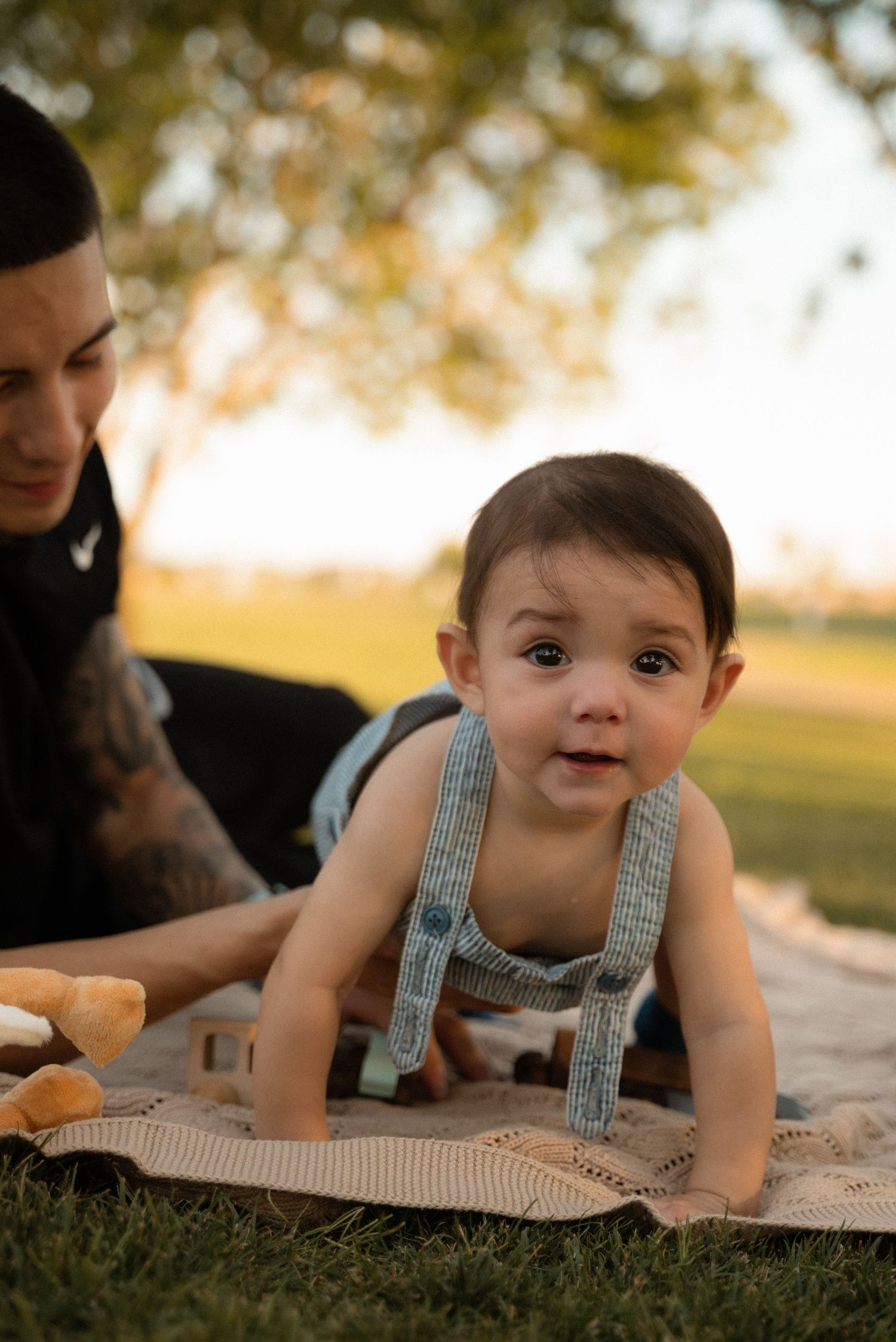 A curious baby in striped overalls crawls on a blanket outdoors at golden hour with an adult nearby.