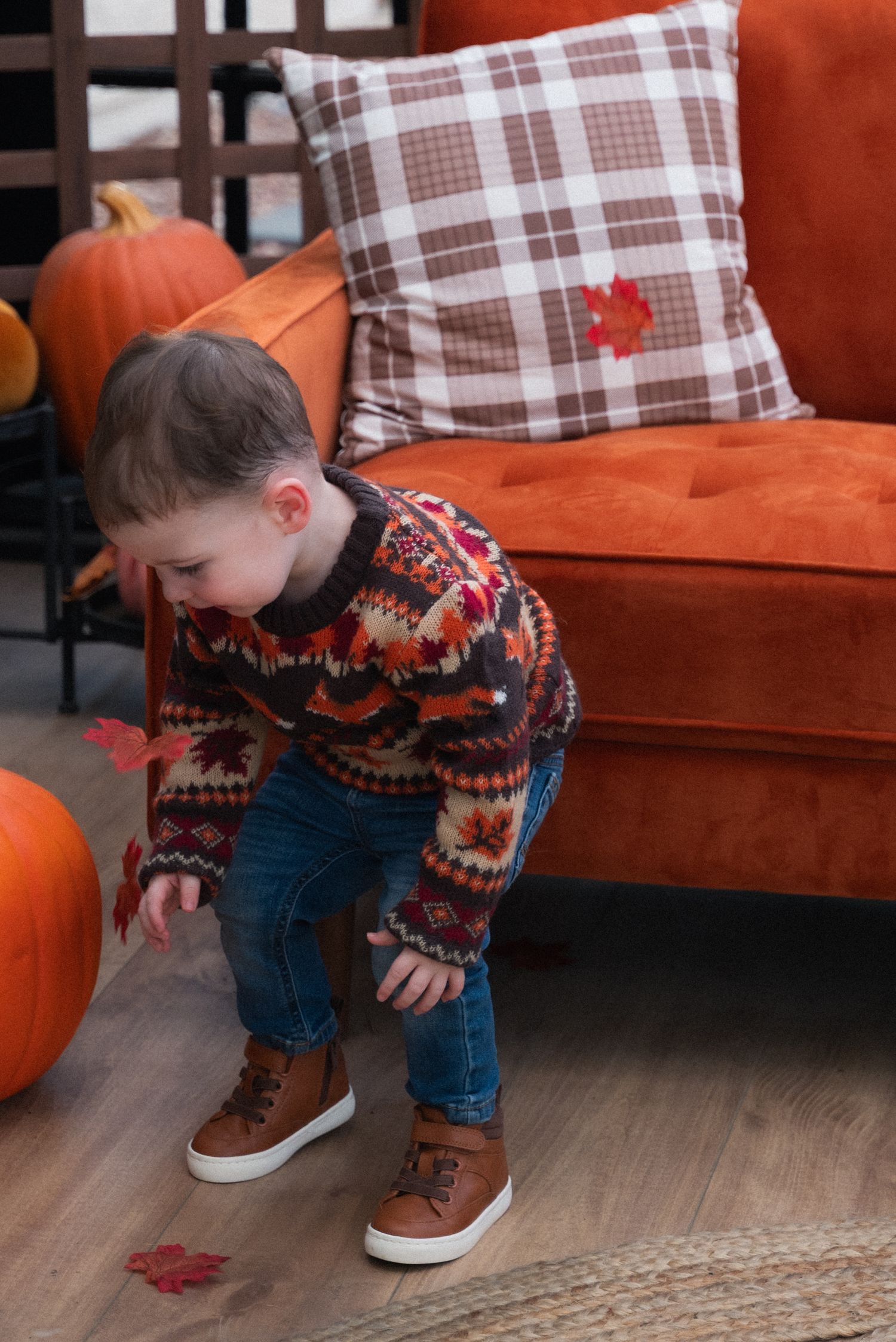 A cozy fall scene with a plaid pillow, orange couch, and pumpkin decor as a small figure bends down to explore.