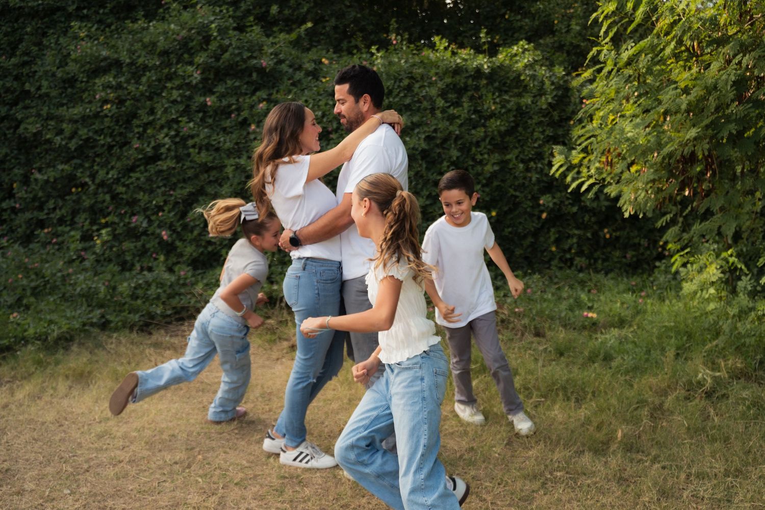A couple in jeans and white shirts dance together in a sunny outdoor setting with green foliage in the background.
