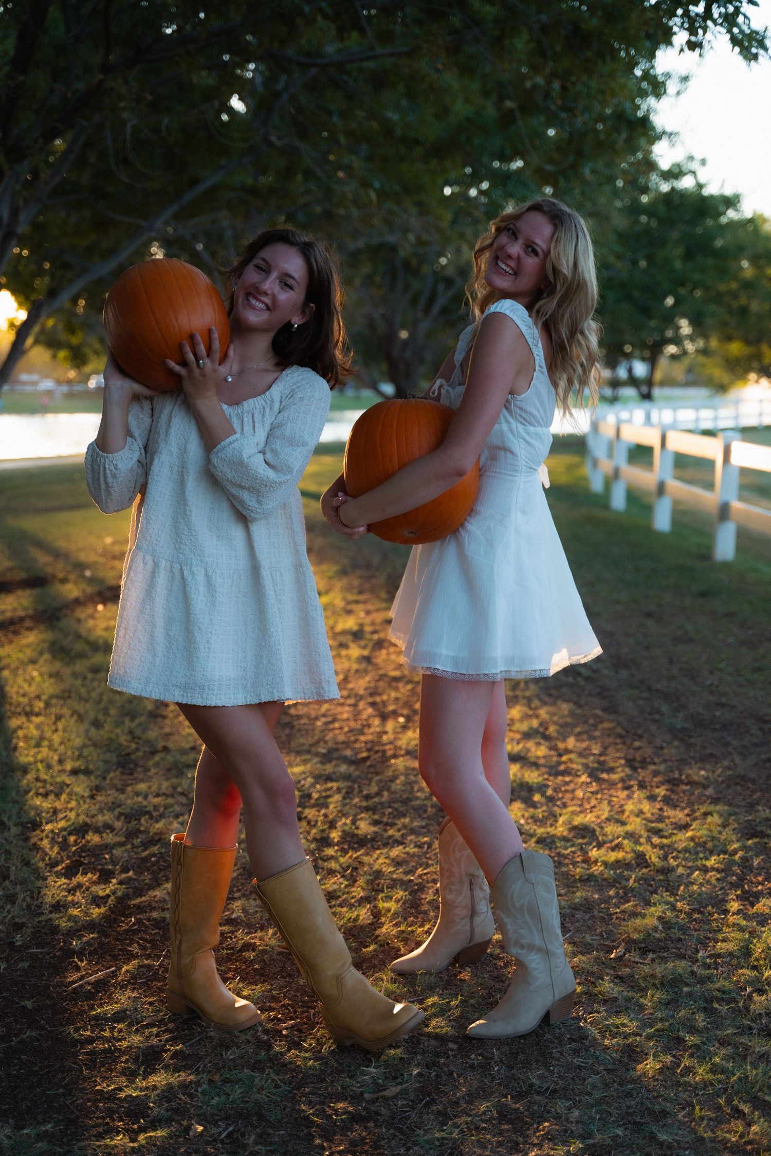 Two people in white dresses and brown boots stand together on fallen autumn leaves during golden hour.