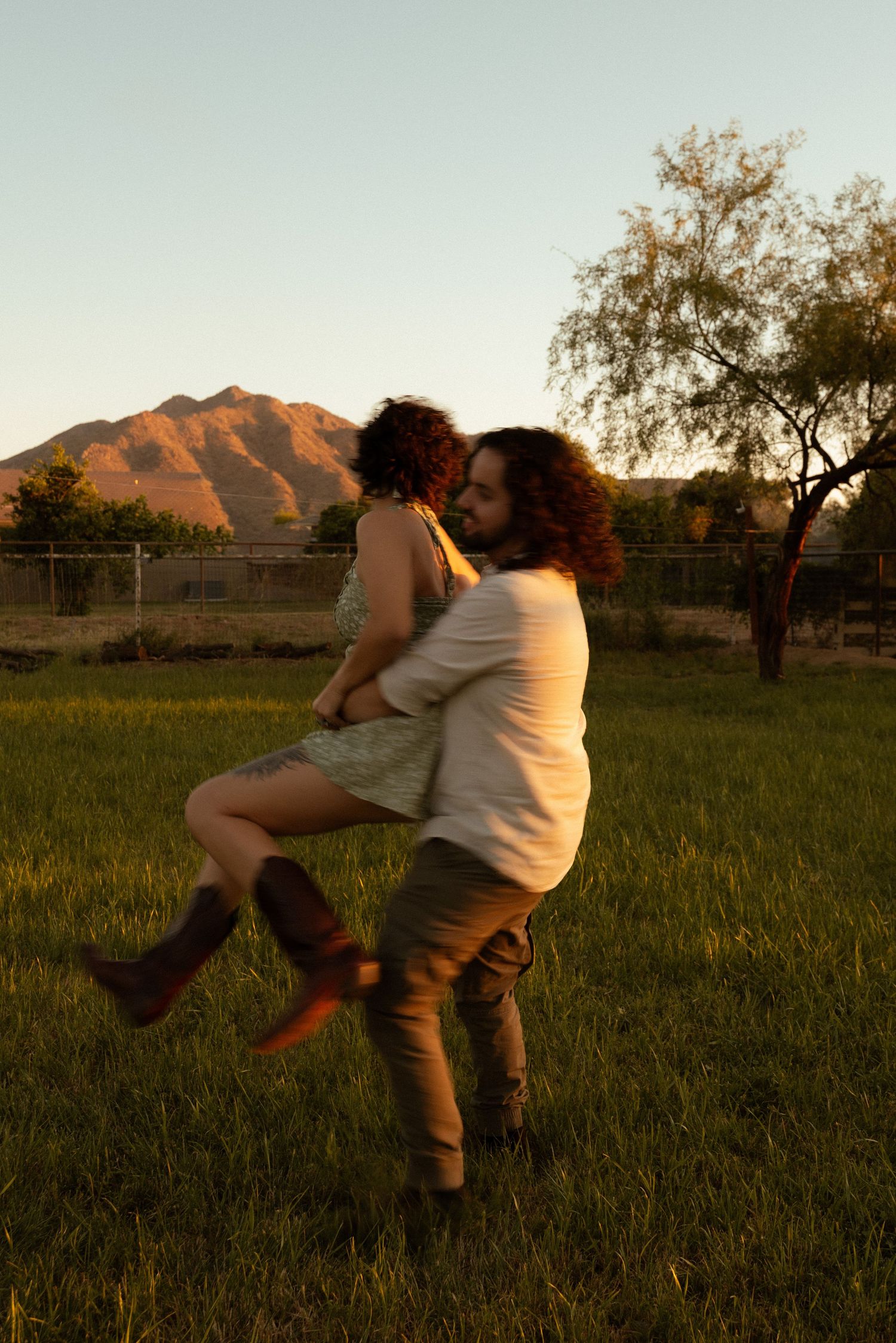 Couple sharing a romantic embrace at golden hour in a green field with a mountain backdrop.