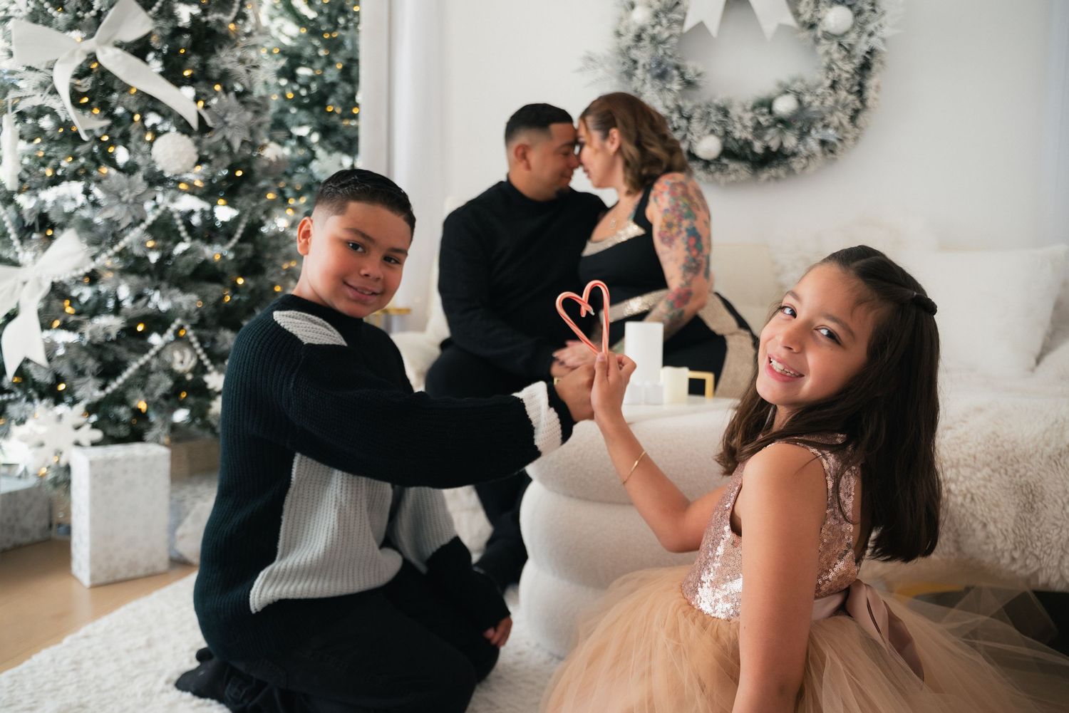 Family Christmas photo with two kids holding candy canes, parents sharing a kiss near a decorated holiday tree.