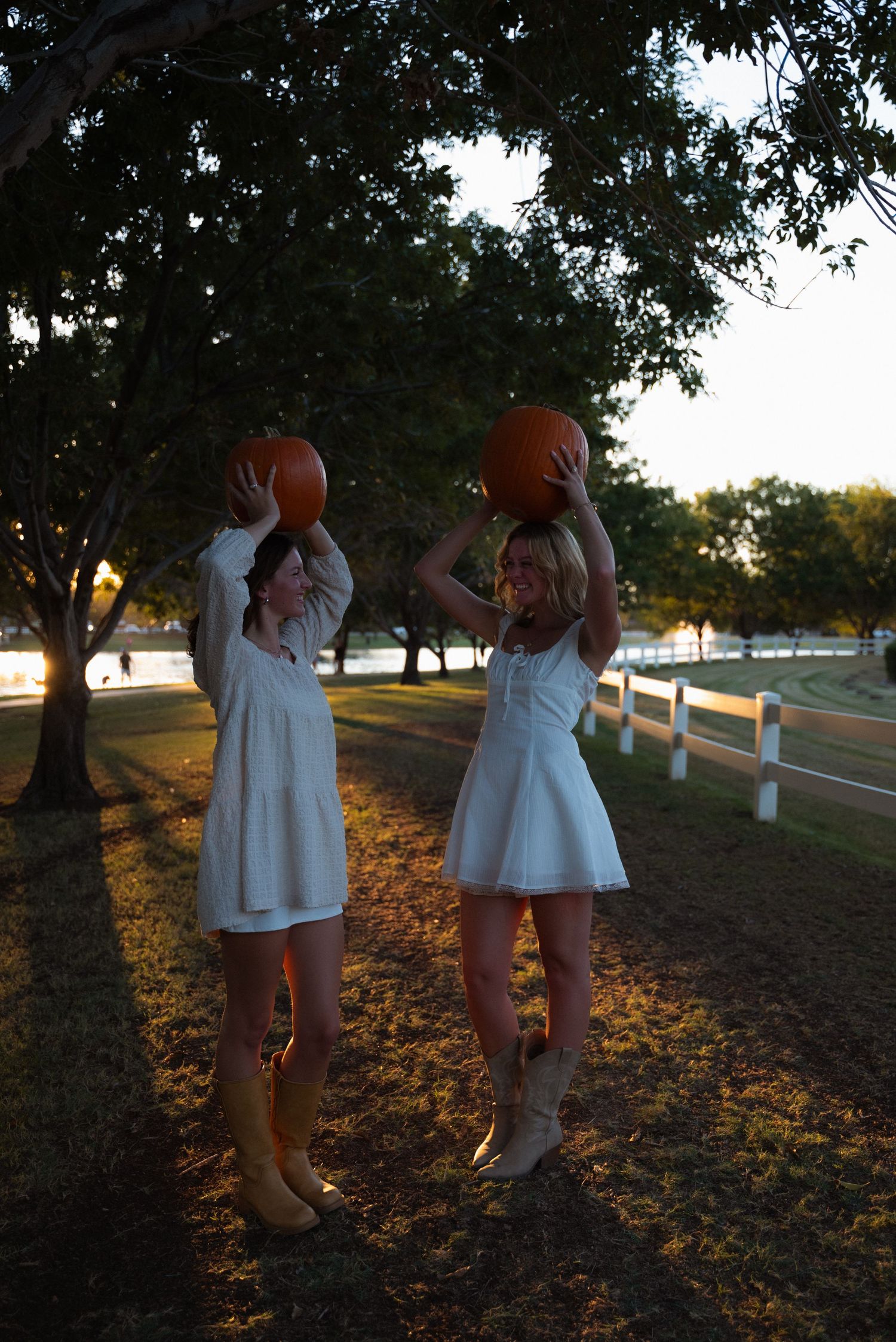 Two people in white dresses pose playfully together at sunset along a fence-lined path with autumn leaves on the ground.