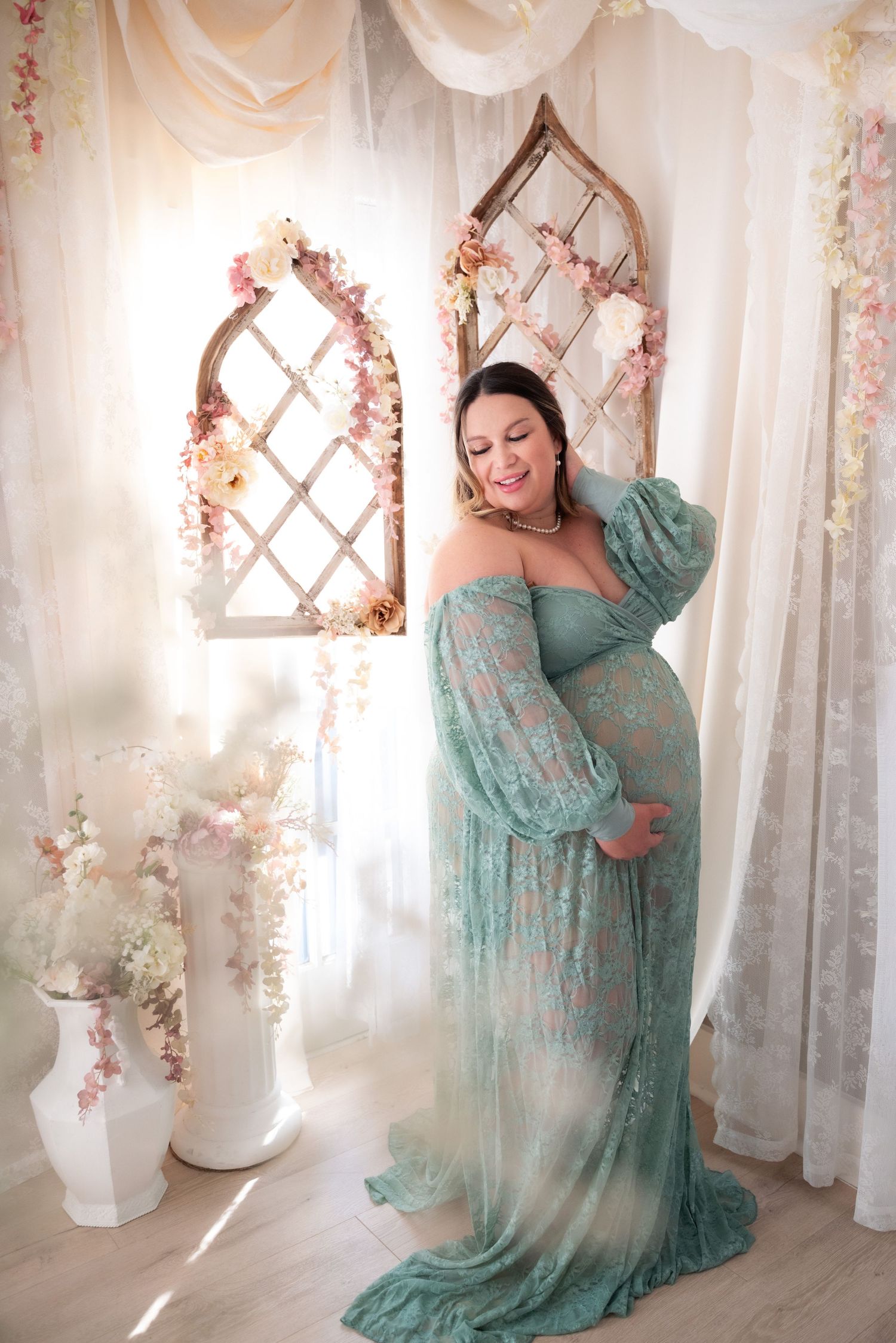 A figure in a flowing mint green dress poses gracefully against white curtains and floral decor in a romantic studio setting.