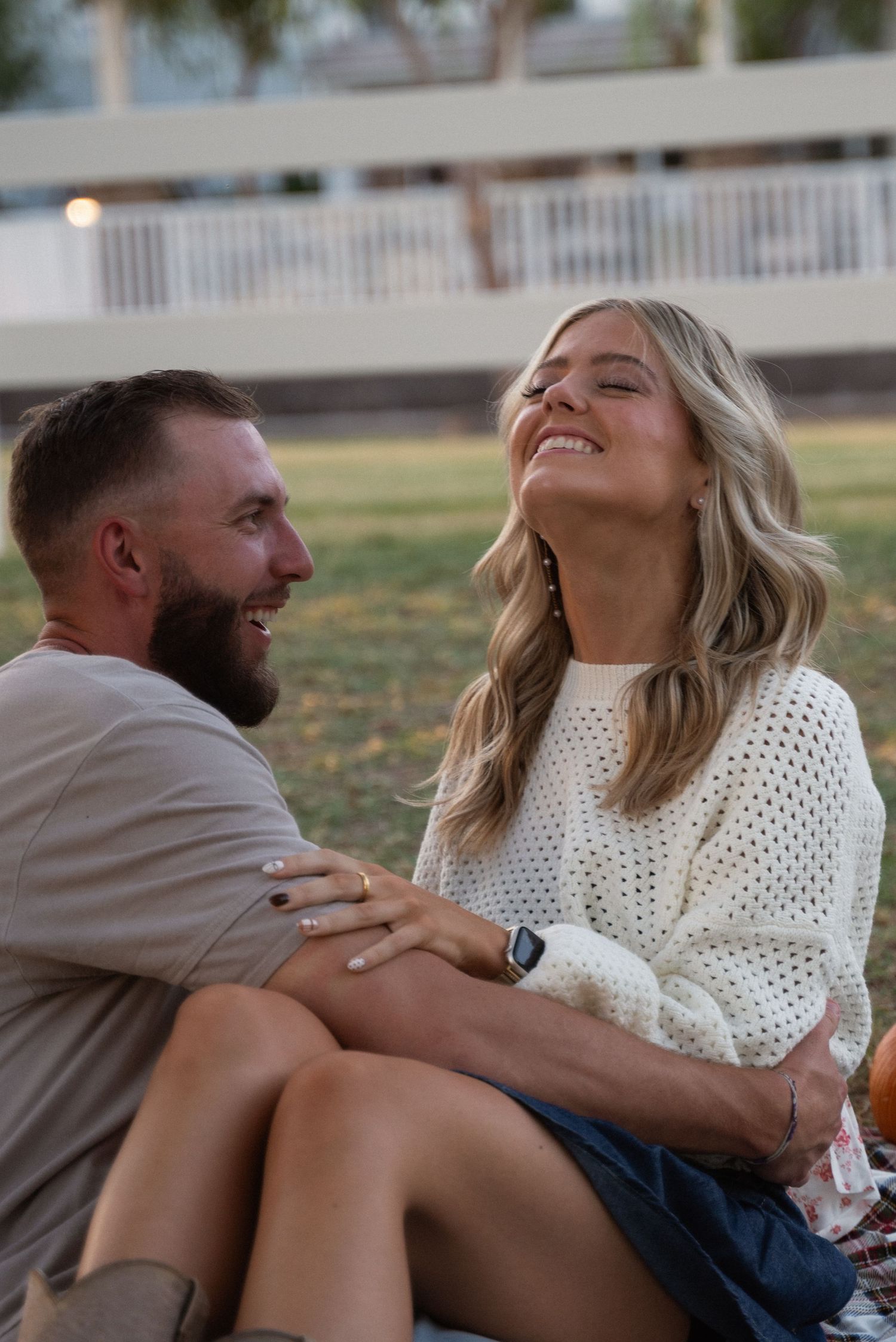 A couple laughs together while sitting on grass during a casual outdoor photoshoot.