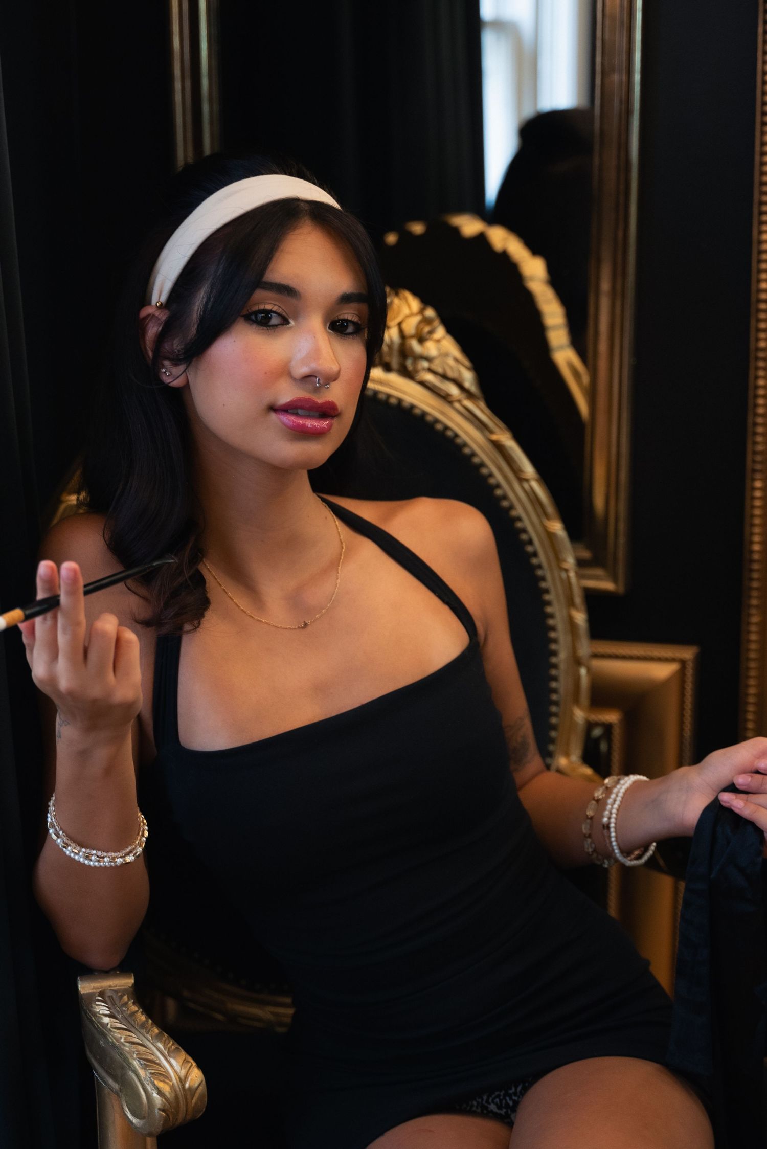 Woman in elegant black dress and white headband poses on ornate vintage chair in moody lighting.