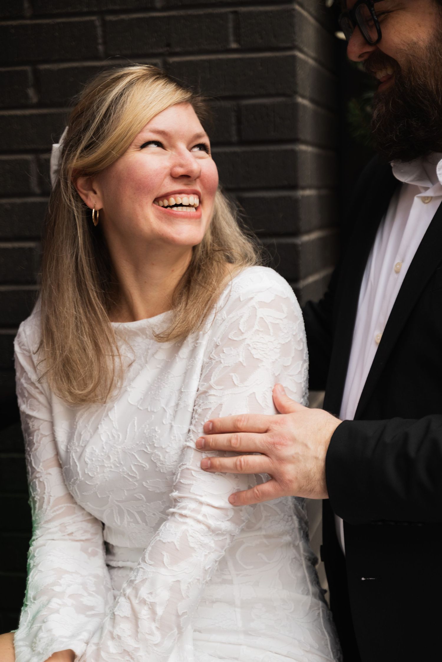 A couple in formal wedding attire sharing a joyful moment against a dark brick wall background.