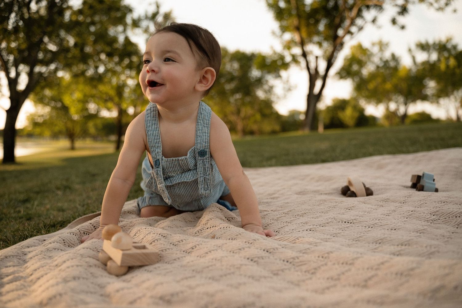 Happy baby in denim overalls laughing on a quilted blanket outdoors in a sunny green park.
