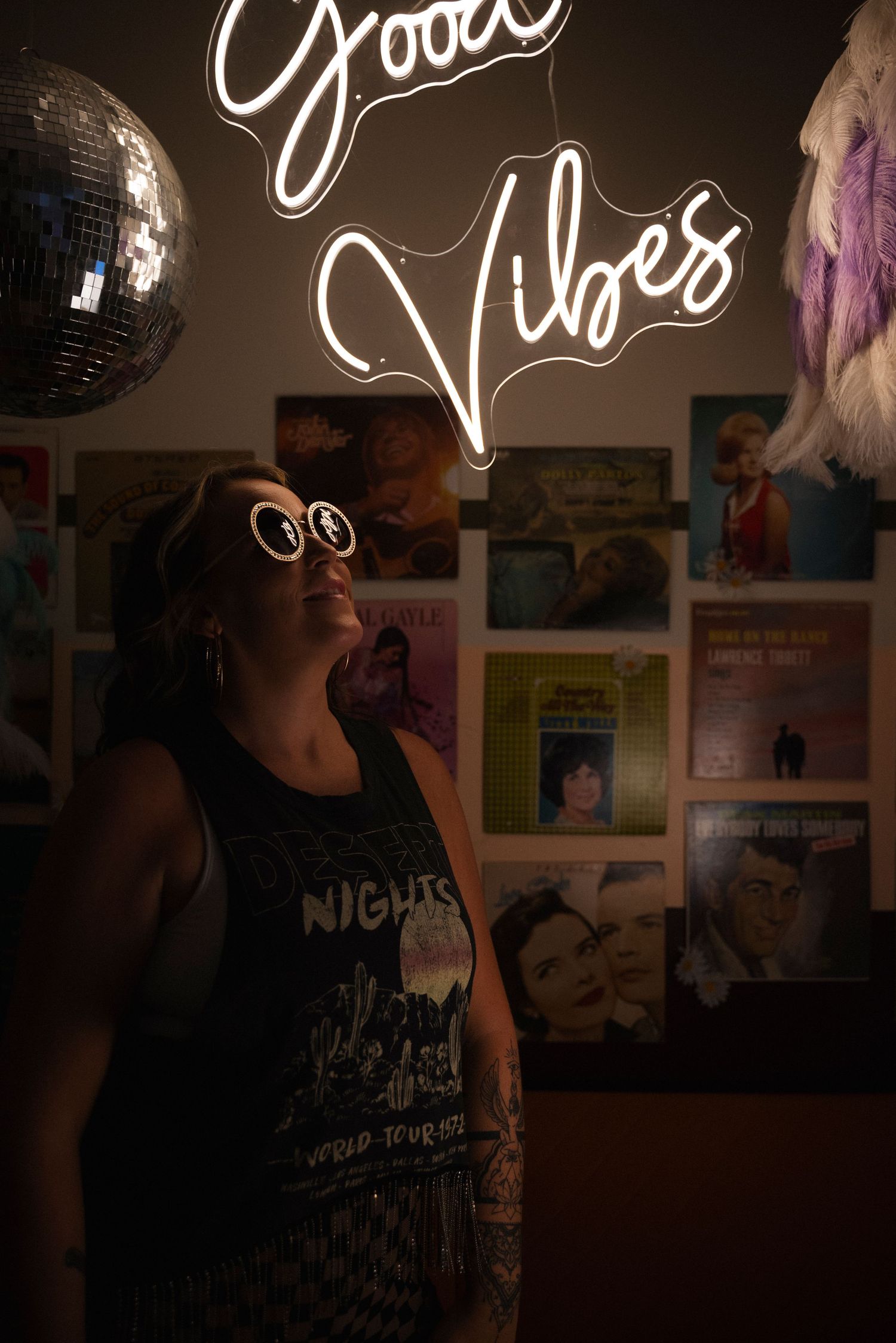 A neon 'Good Vibes' sign glows above a vintage wall collage with a disco ball in a dimly lit room.