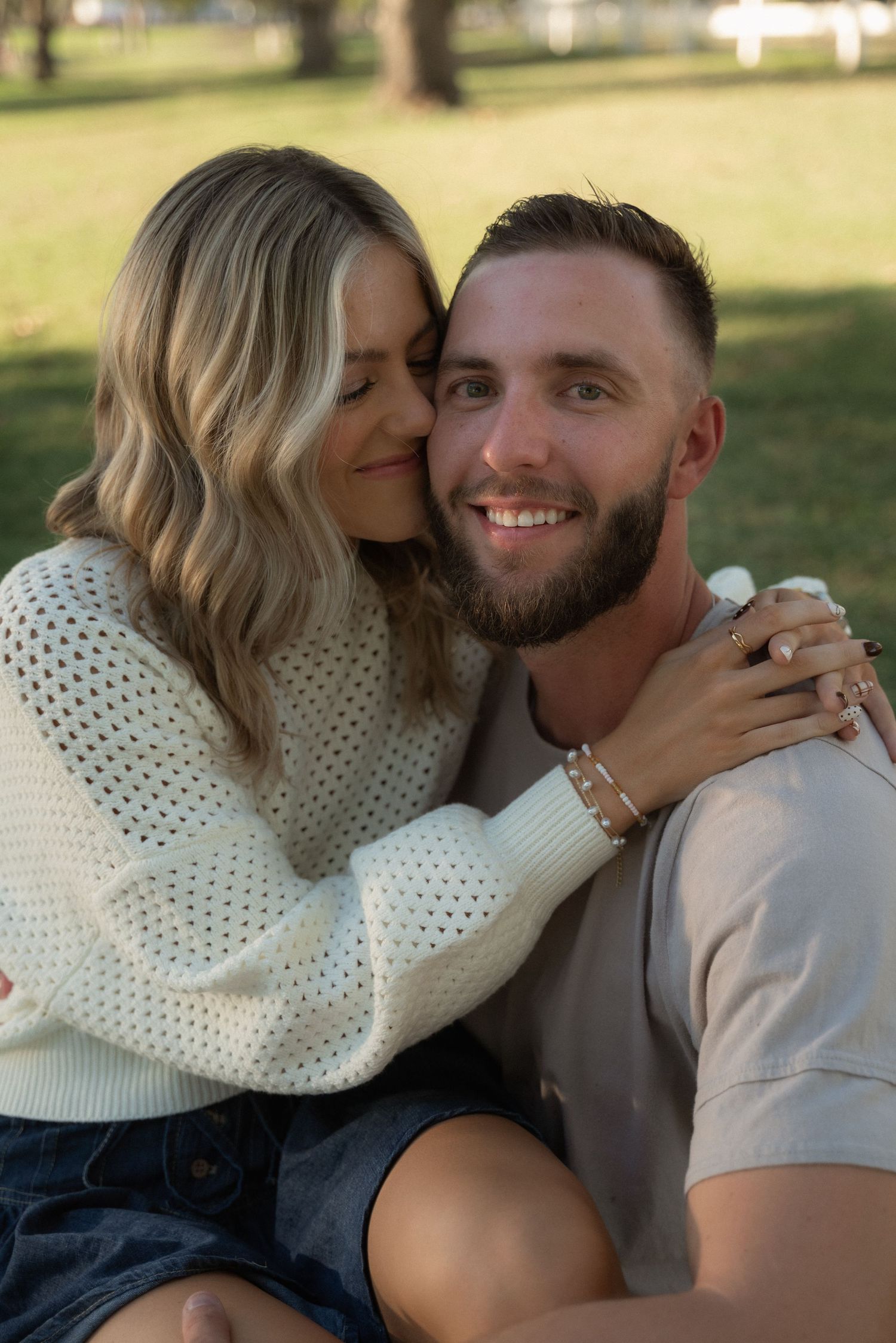 A romantic couple shares an intimate moment outdoors, with her wearing a white knit sweater and him in a gray t-shirt.