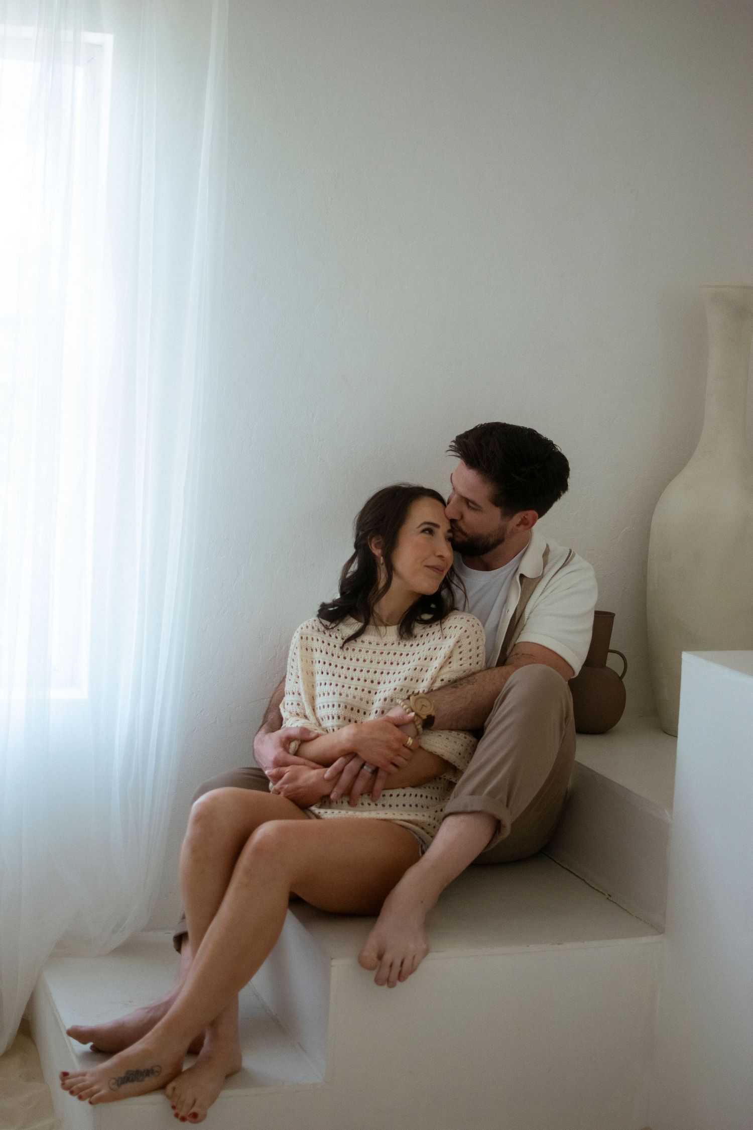 Intimate couple sharing tender moments on white windowsill in minimalist setting with soft natural lighting.