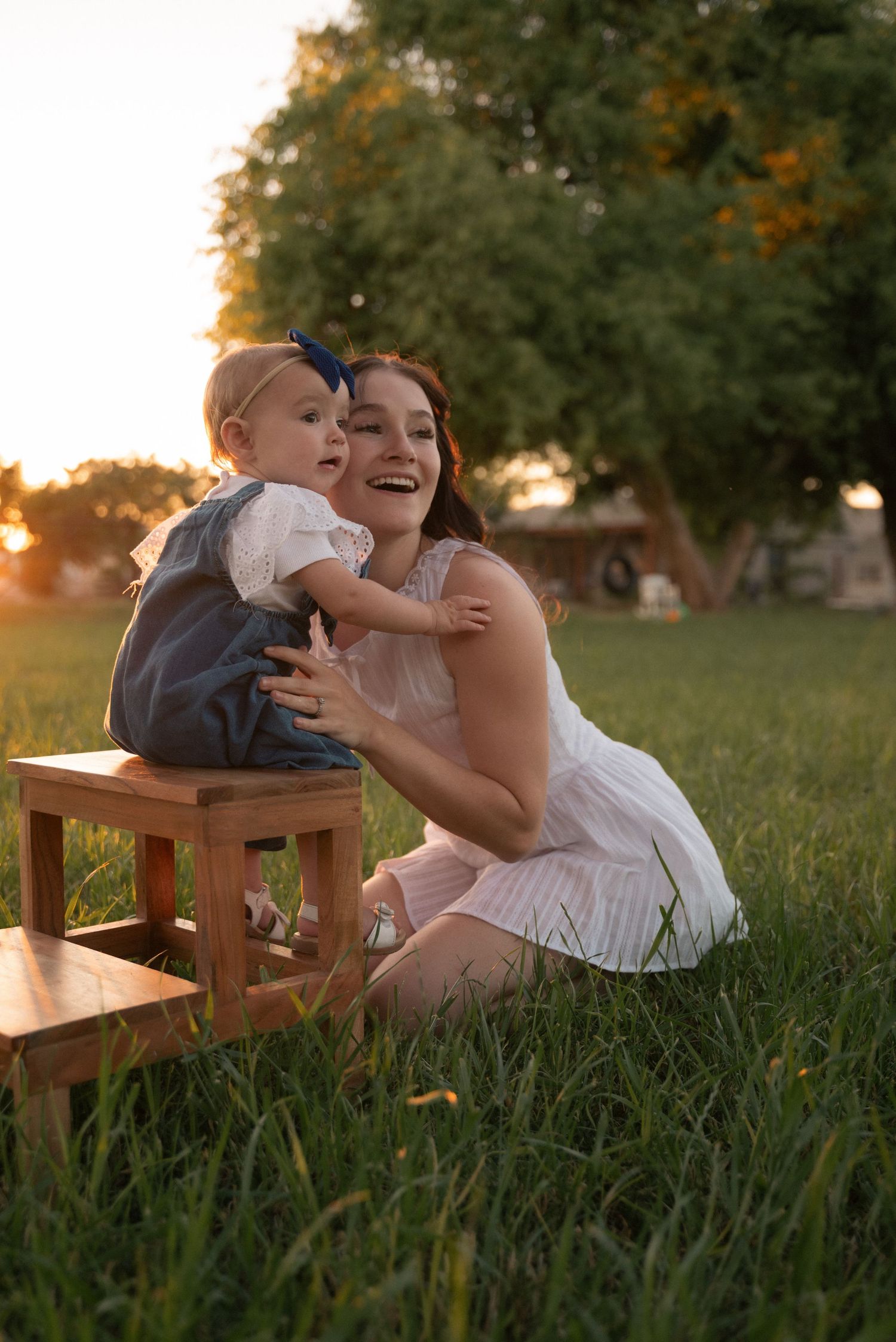 A joyful mother in a white dress hugs her baby on a wooden stool in a sunlit green field at golden hour.