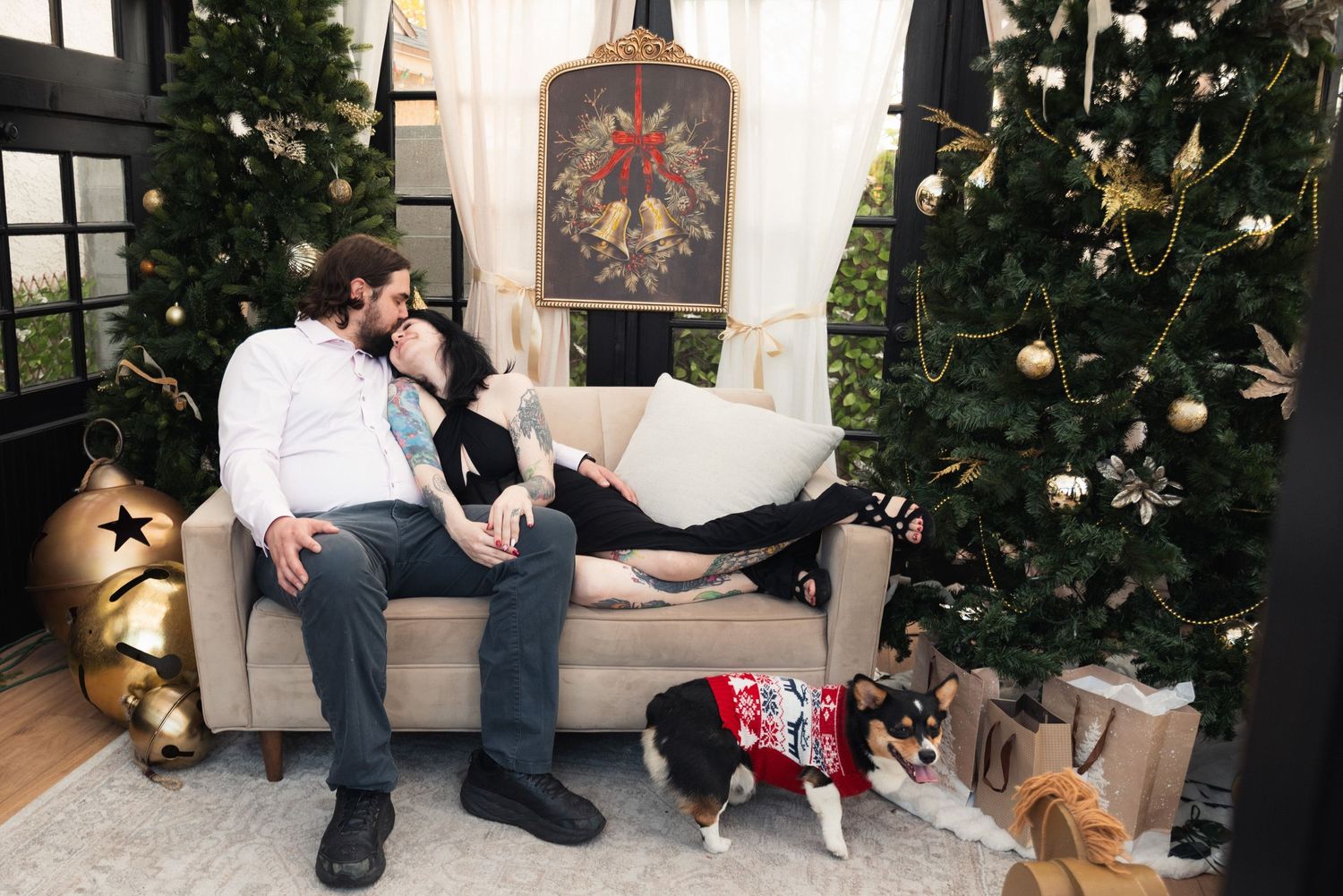 Couple relaxing on a beige sofa surrounded by decorated Christmas trees with a dog in a holiday sweater nearby.