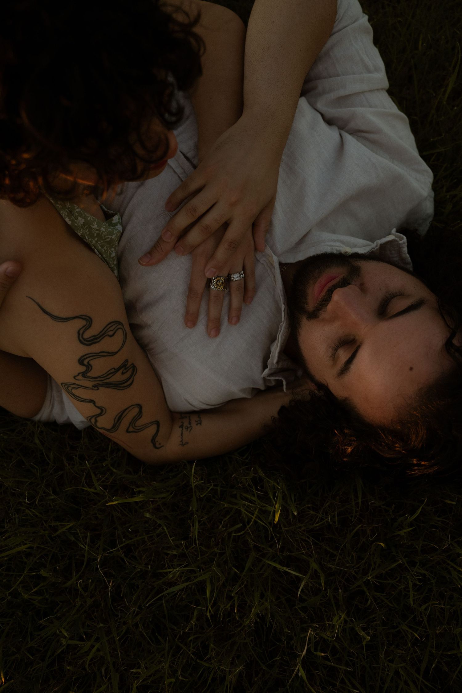 A couple lies on grass, woman with snake tattoo rests hand on man's chest wearing a ring, eyes closed.
