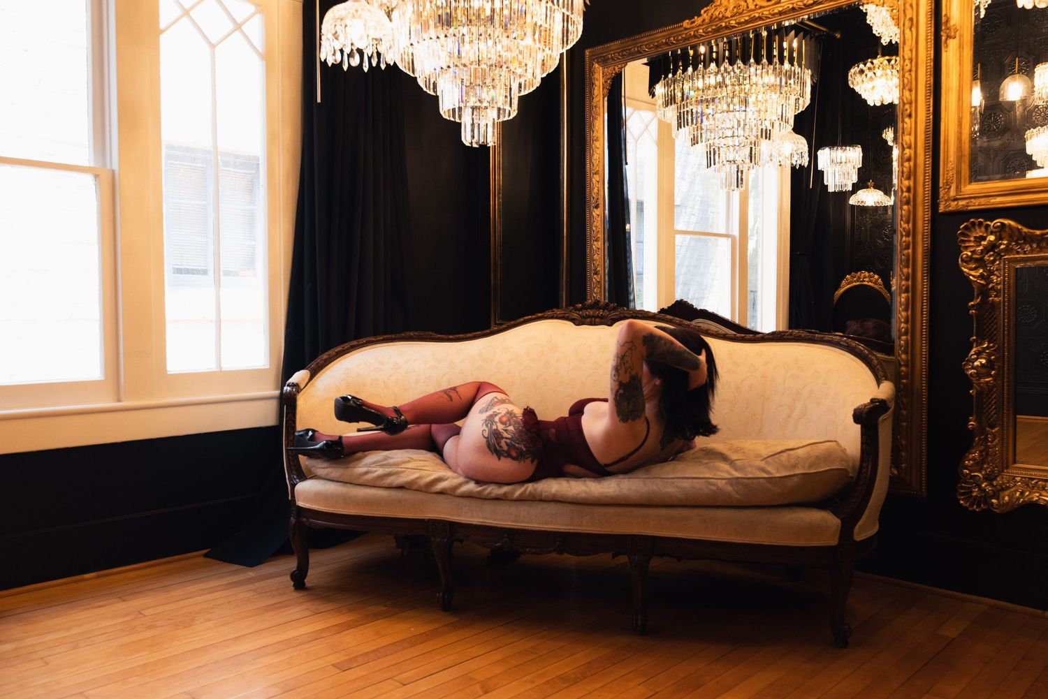 Woman in red lingerie lounging on ornate vintage sofa under crystal chandeliers in elegant dark room.
