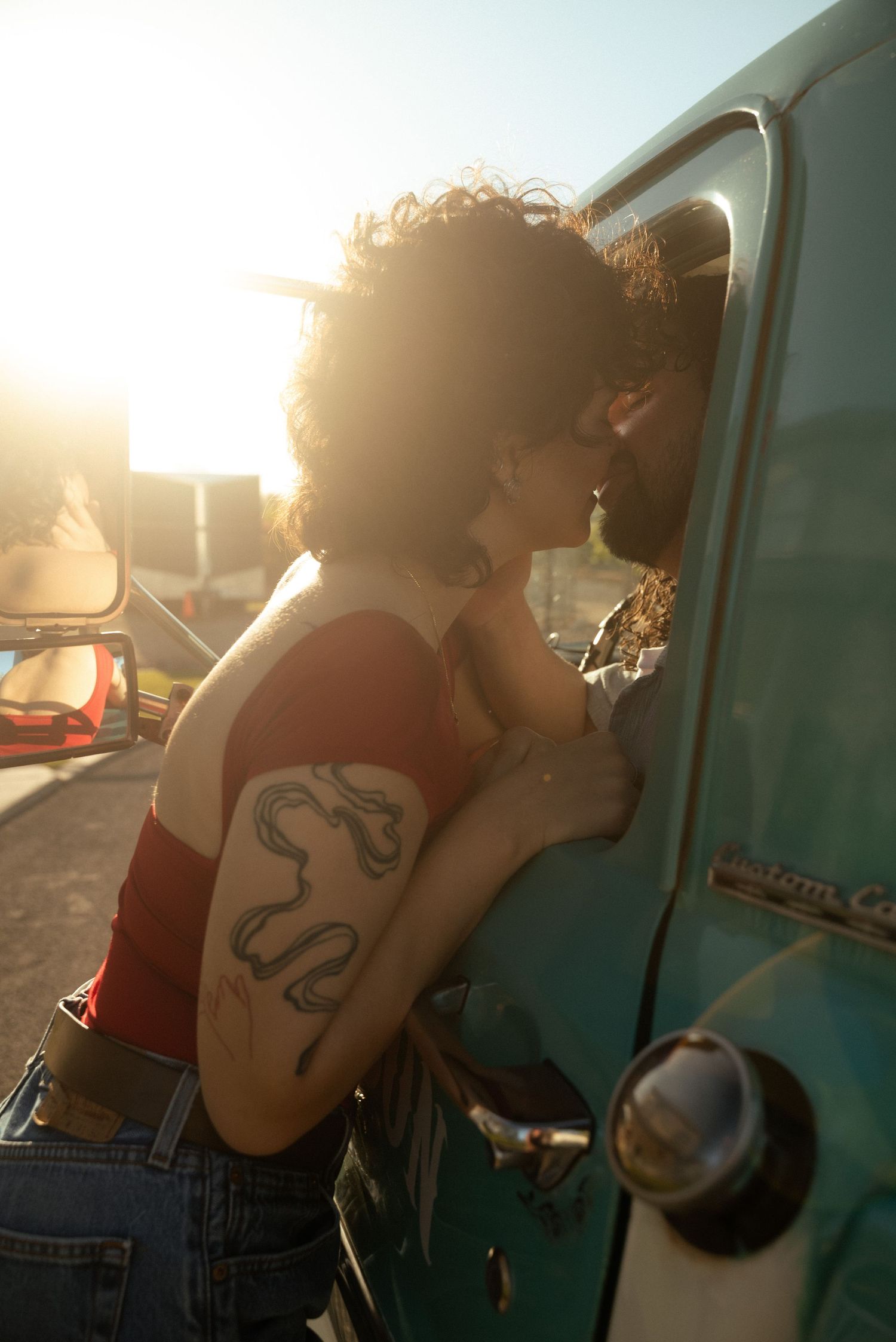 A tattooed woman leans into a vintage teal truck window sharing a kiss at golden hour sunset.