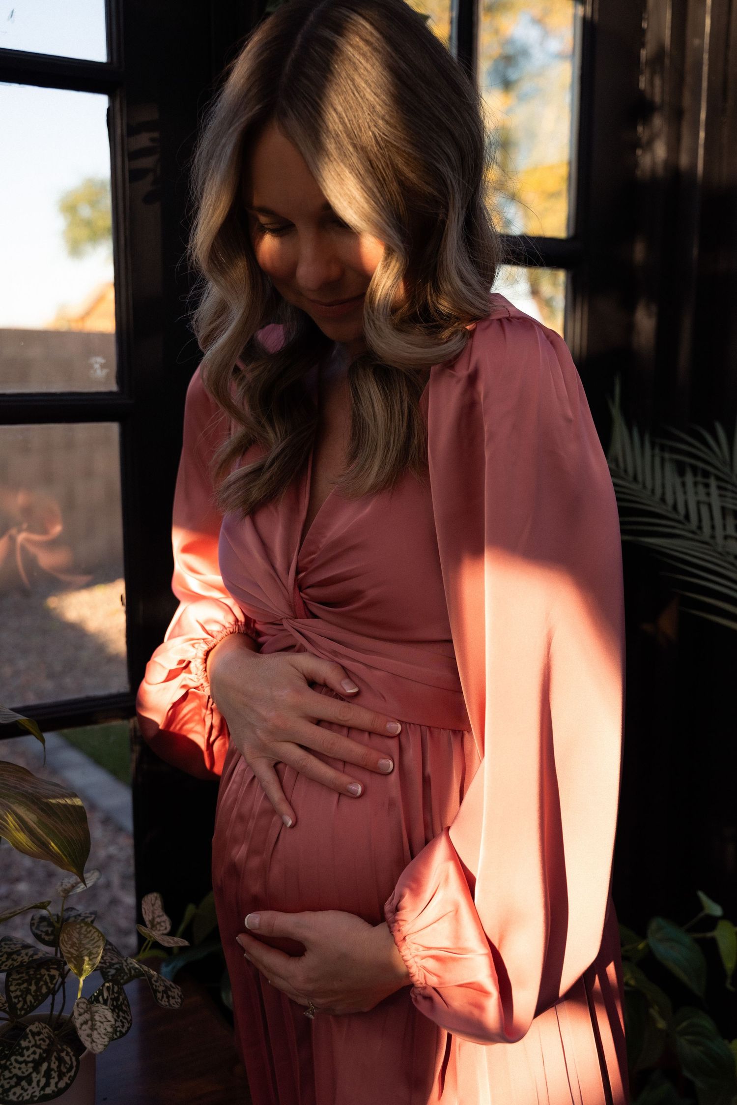A series of photos showing a pregnant person in a pink dress cradling their belly in warm sunlight.