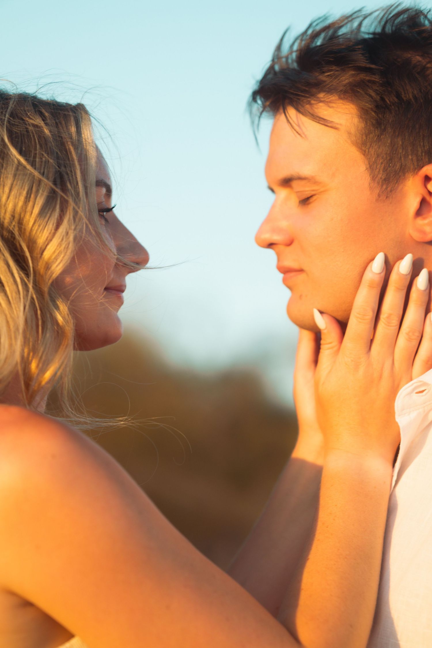 A young couple shares a romantic moment at golden hour, with a woman tenderly holding a man's face close to hers.
