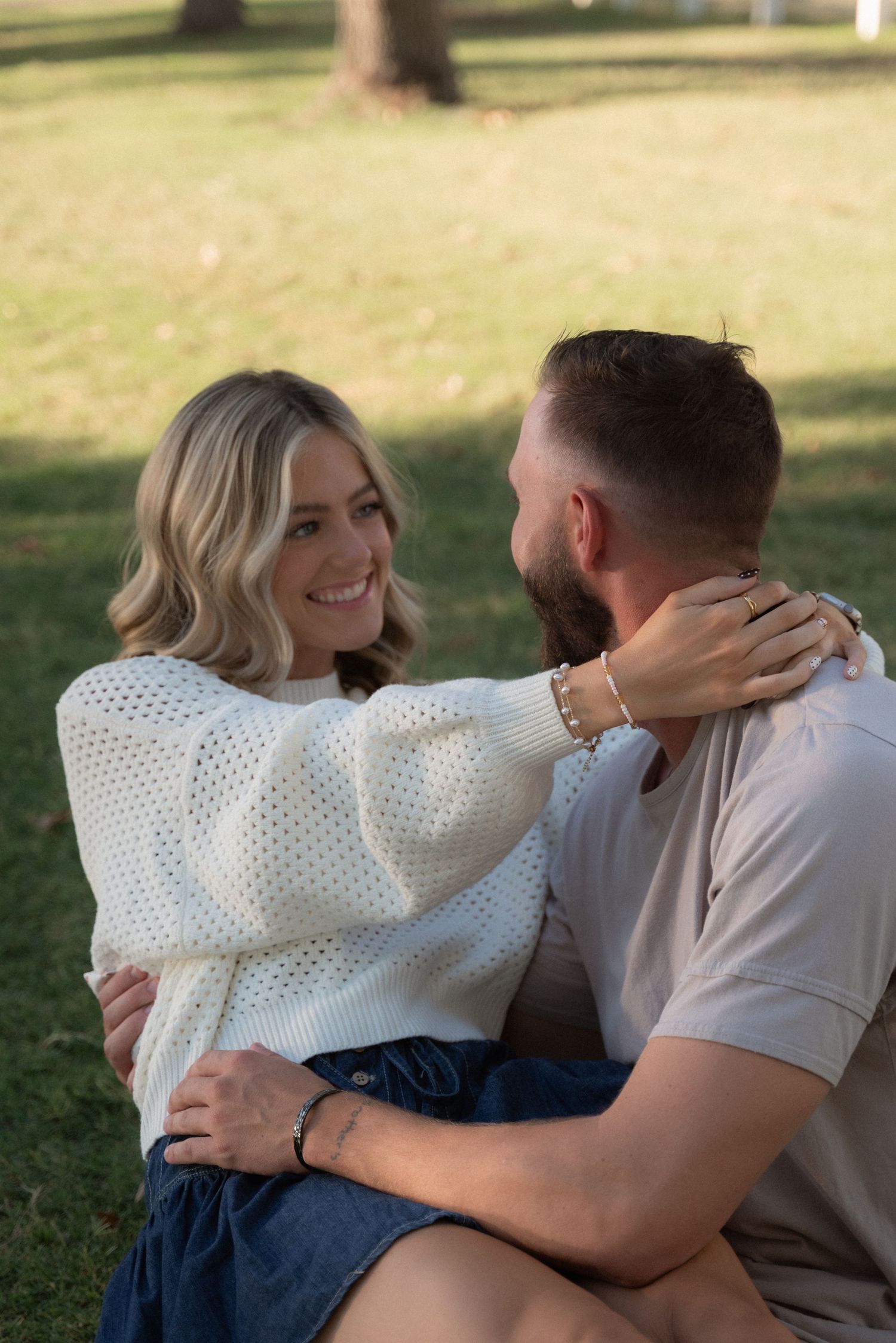 A couple in casual attire sharing an intimate embrace while sitting outdoors on grass in natural lighting.