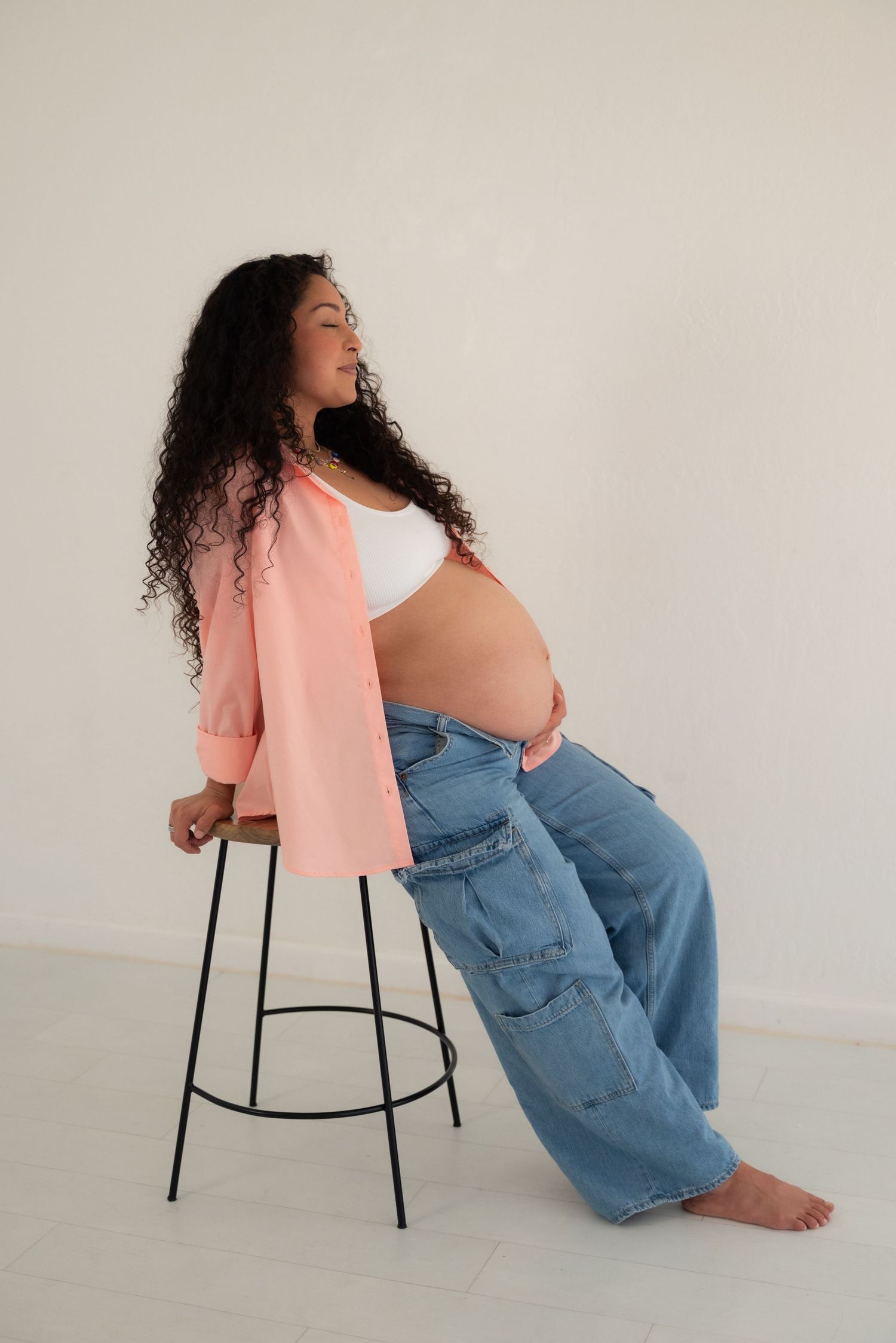 A person in a peach blazer and blue jeans sits sideways on a black chair against a white backdrop.