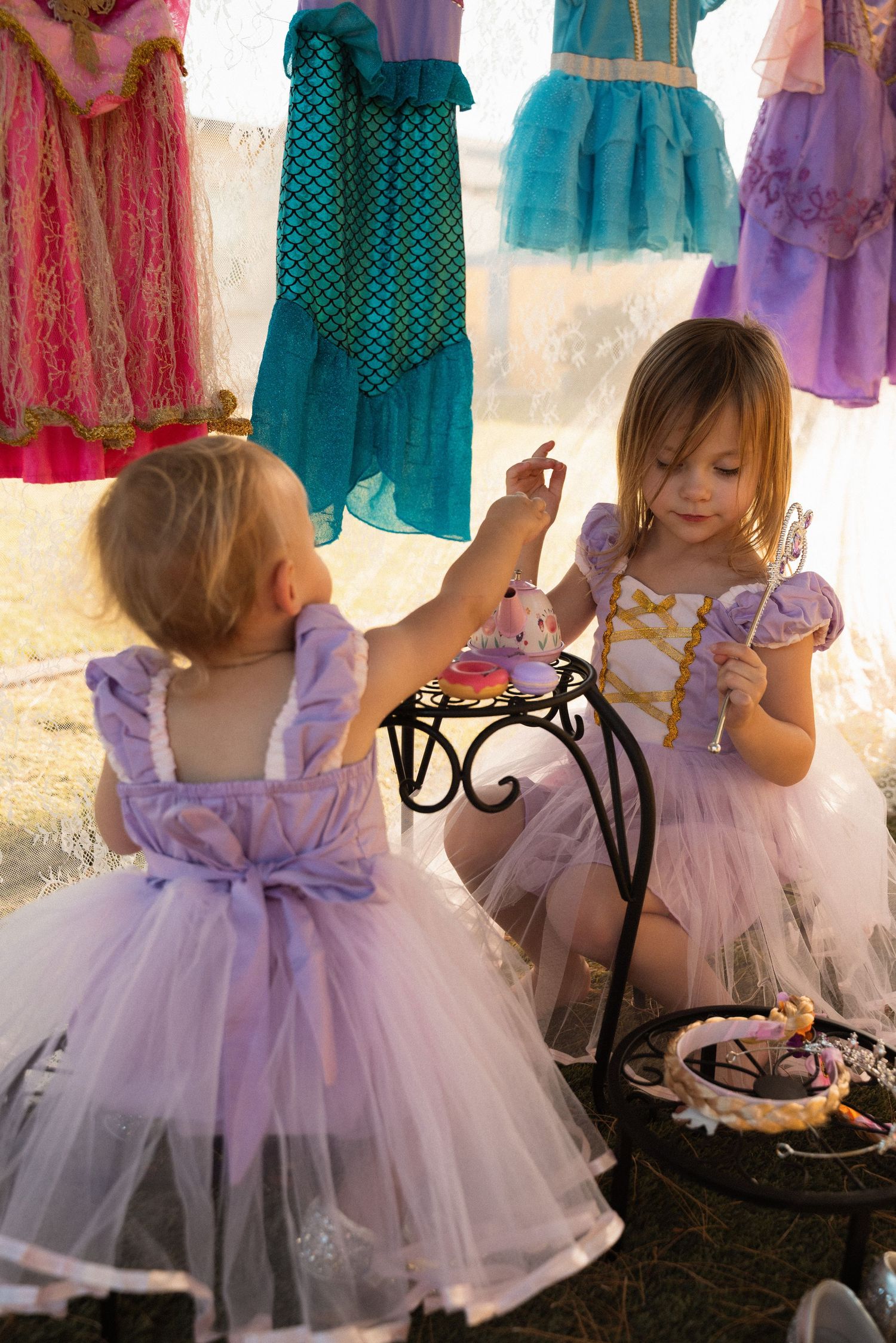 Two little girls in purple princess dresses play at a tea party table with colorful costumes hanging in the background.
