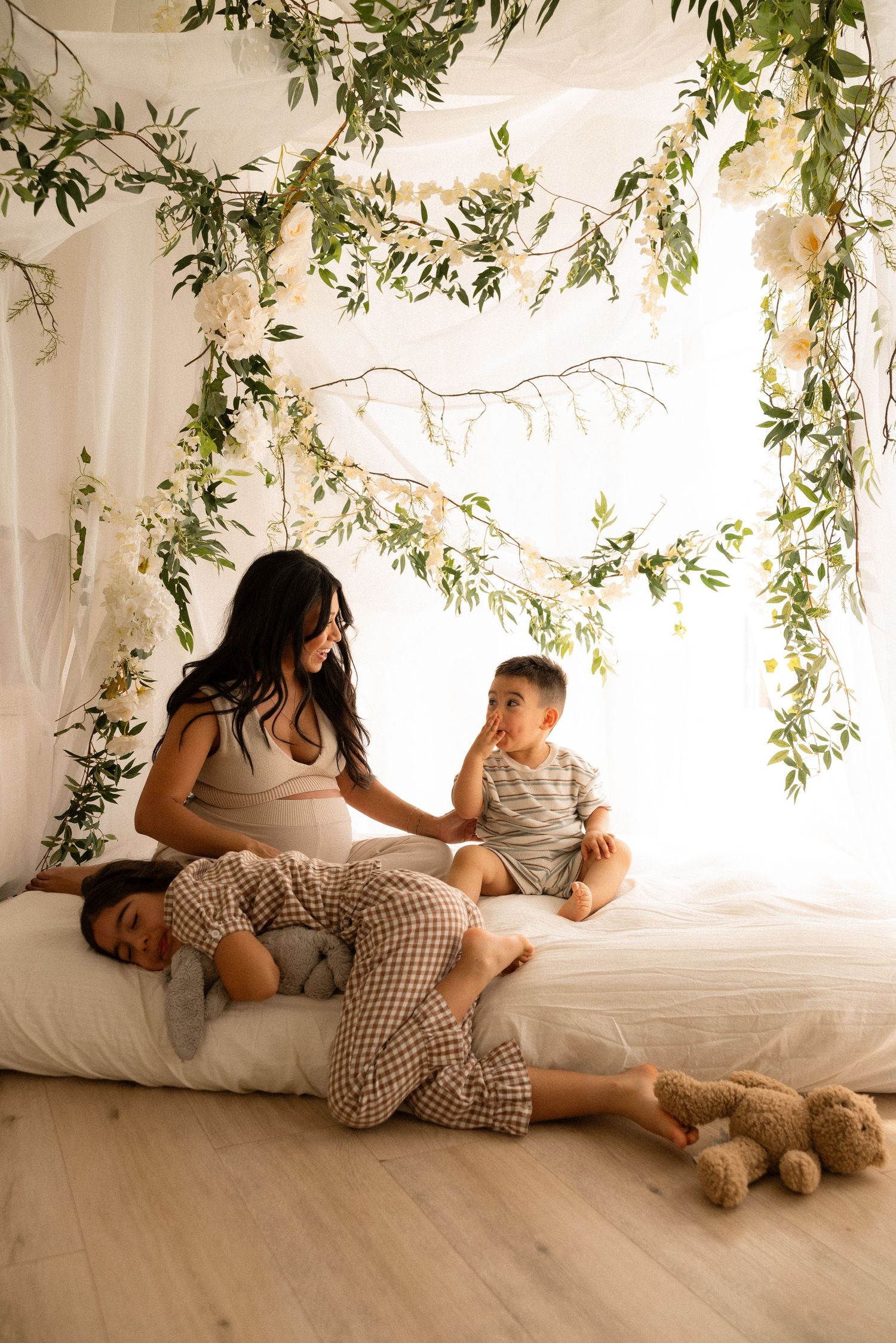 Pregnant woman sits on white bed with two children under floral greenery canopy in bright airy room.