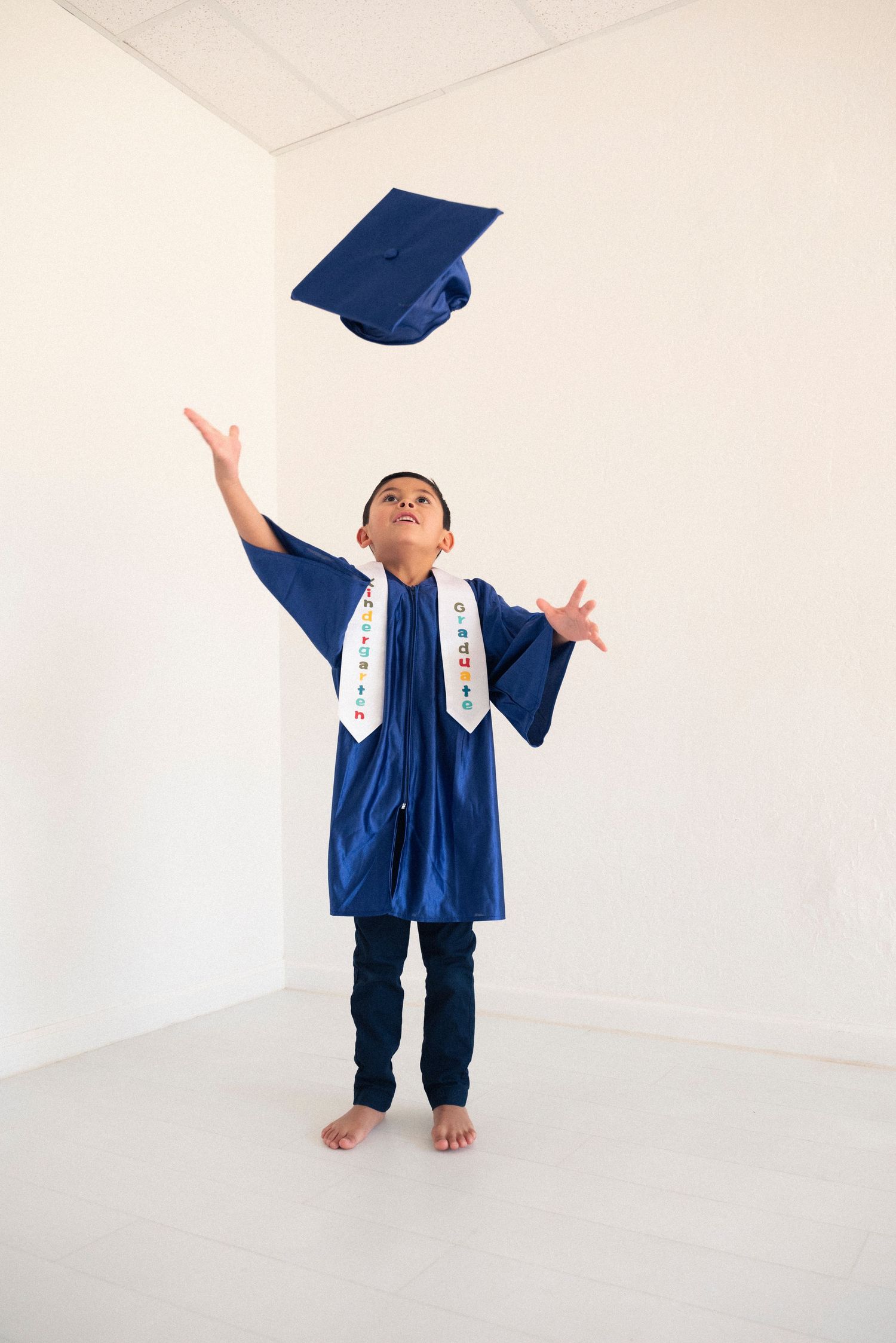 A graduate in a navy blue cap and gown throws their graduation cap in the air during a photo shoot against a white wall.