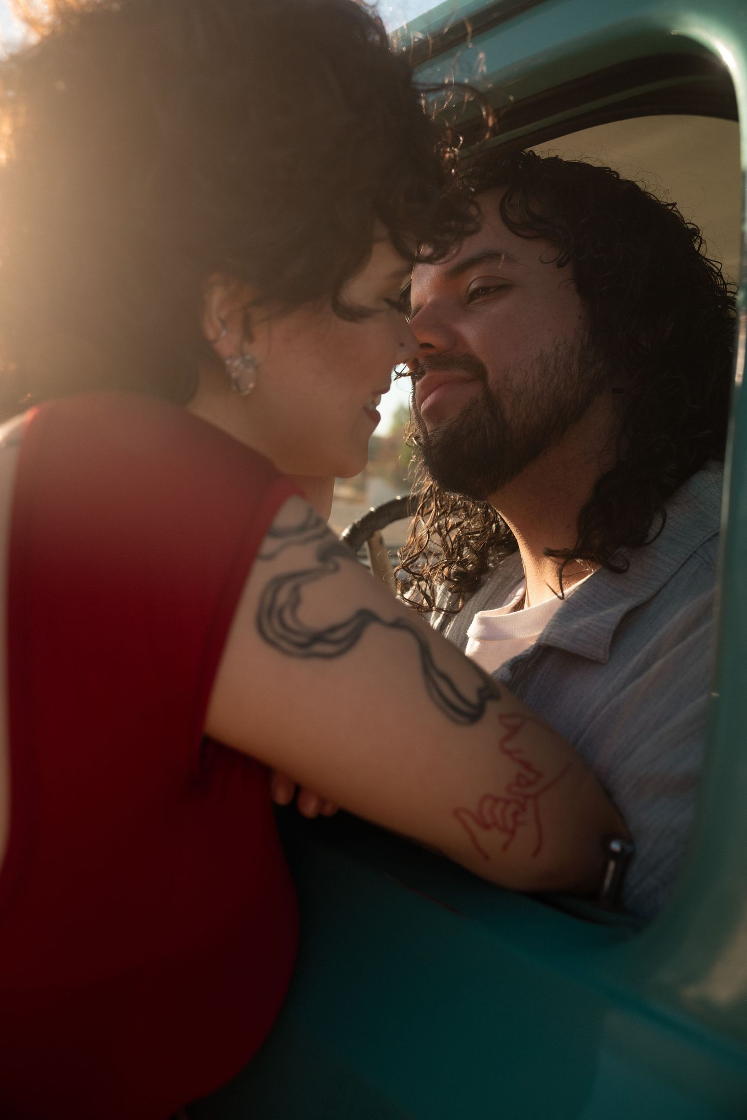 Couple sharing a romantic moment in a vintage teal truck, woman with tattoos in red dress, golden hour lighting.