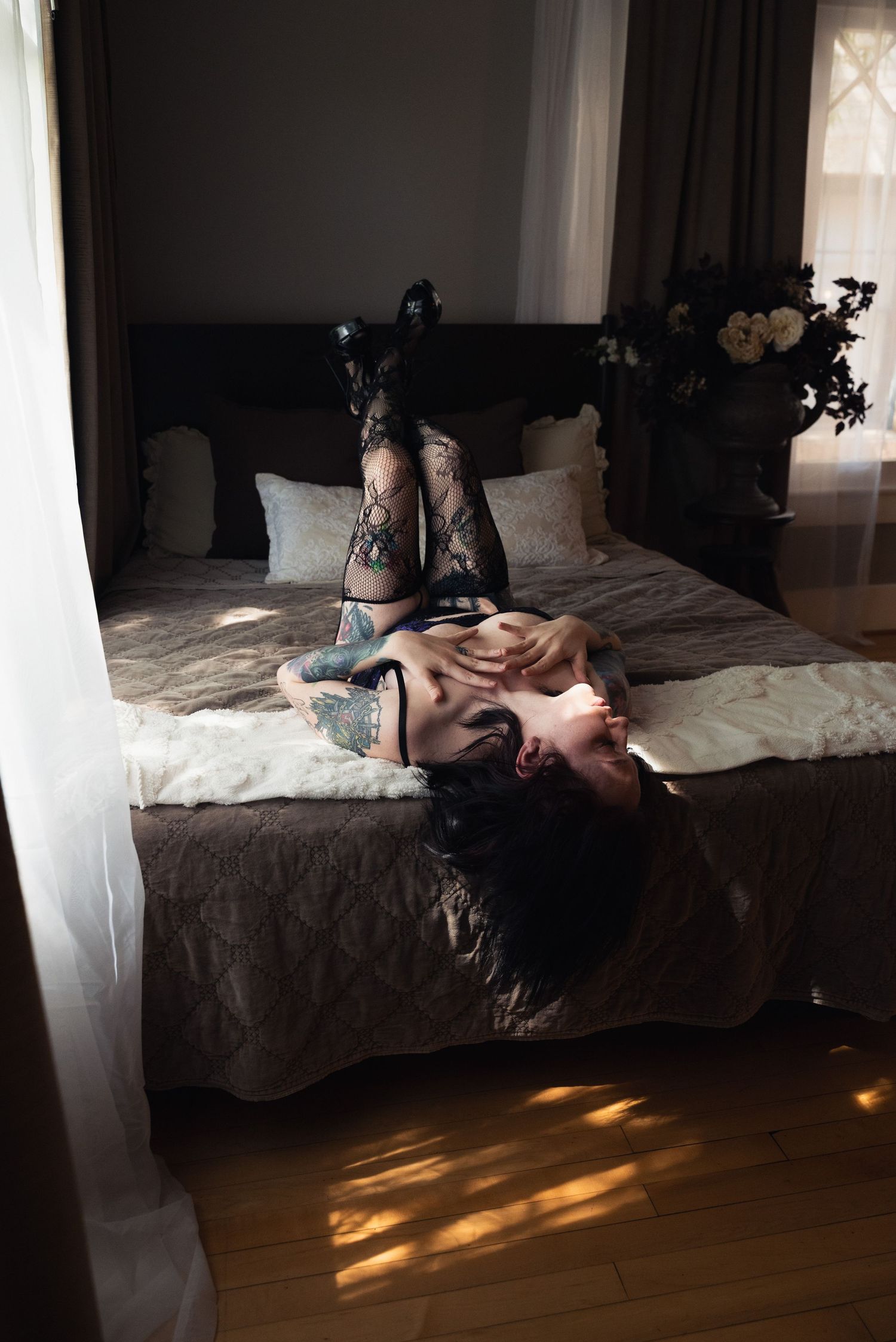 Woman in black lace lingerie and stockings posing on a bed in a softly lit bedroom.