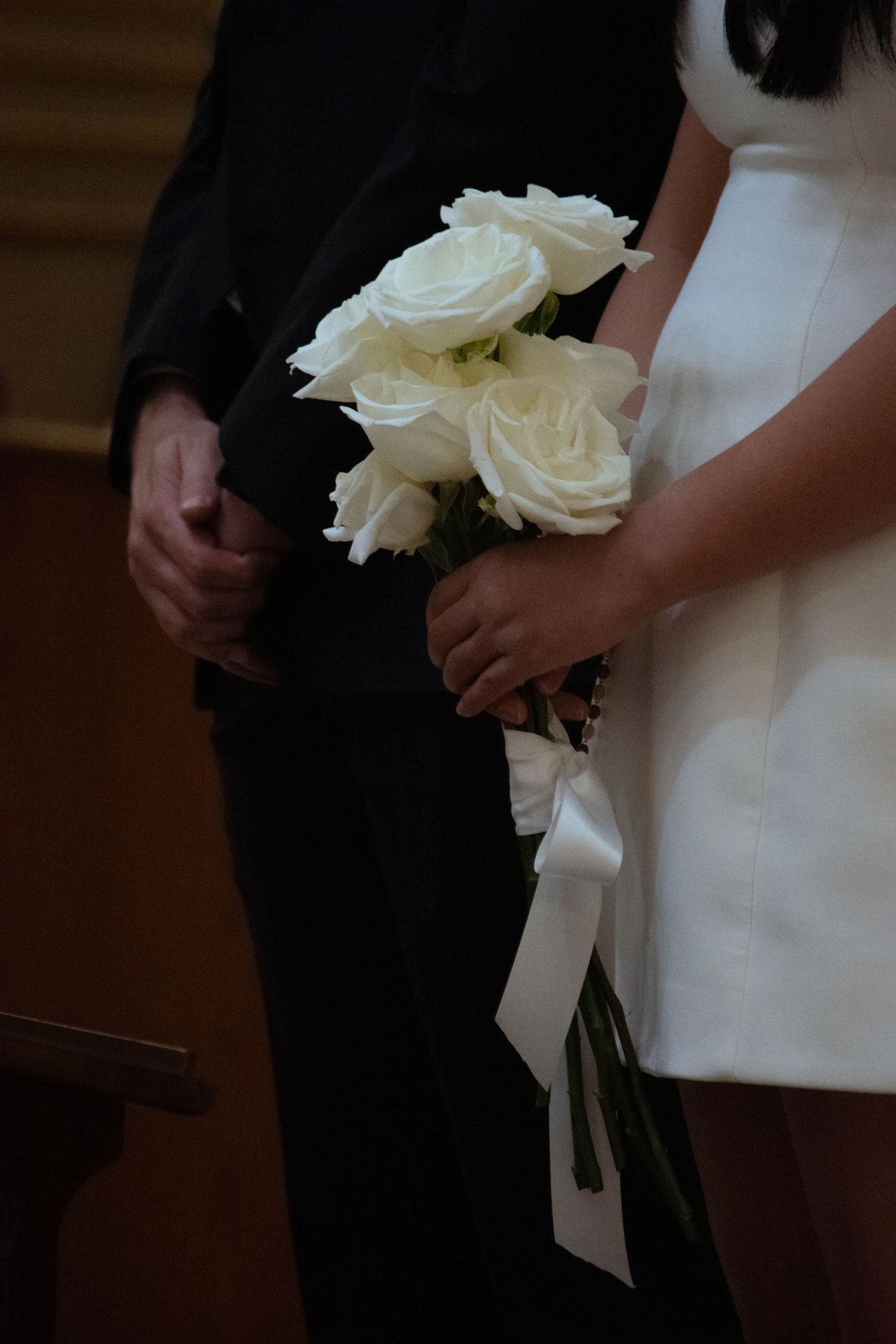 A person in formal attire holds a white rose bouquet with a flowing satin ribbon against a dark background.